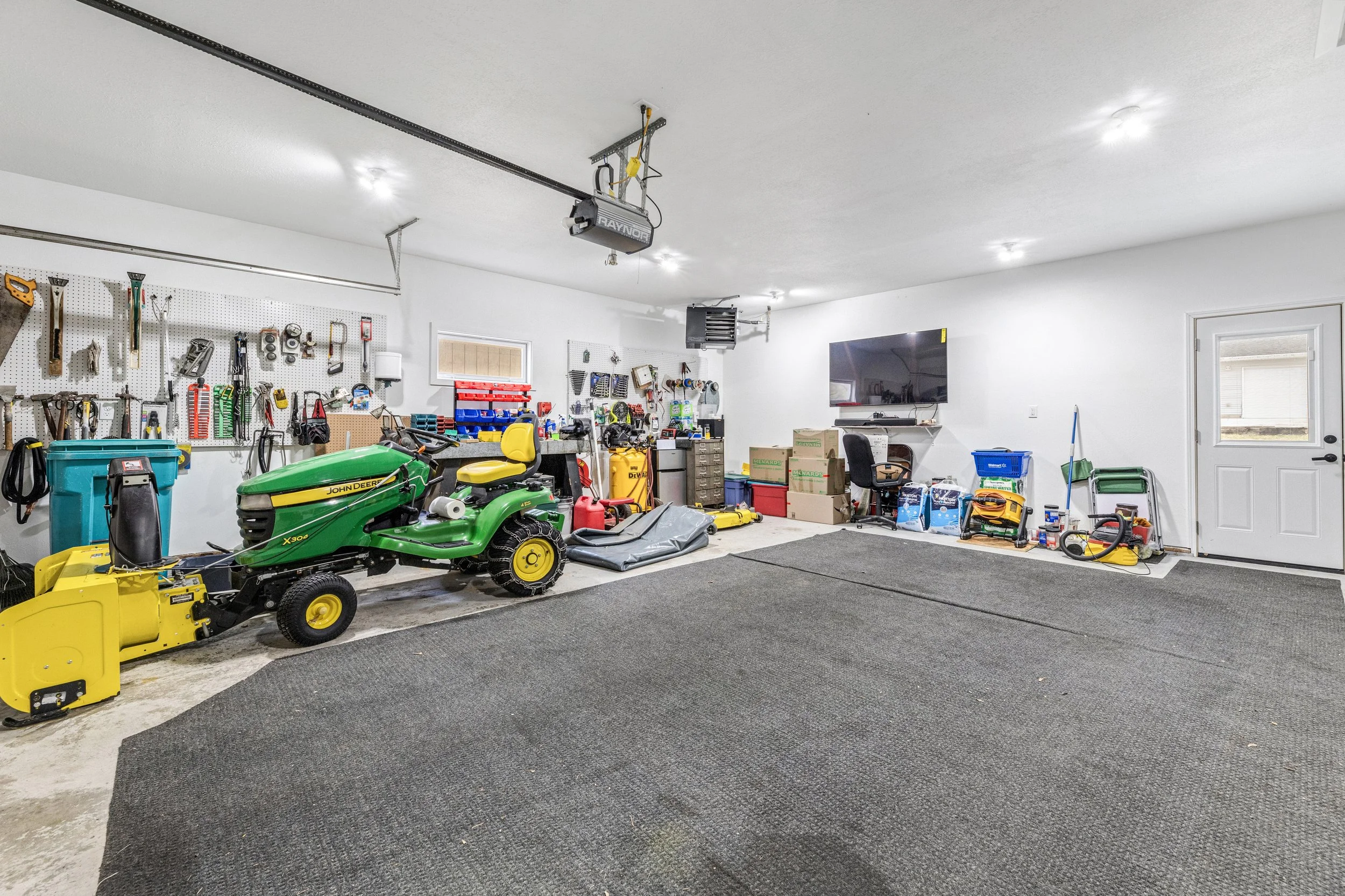 A spacious garage with various tools hanging on a pegboard wall, a John Deere riding mower, and boxes of supplies. The ceiling has a garage door opener, and there are black and gray mats on the floor.