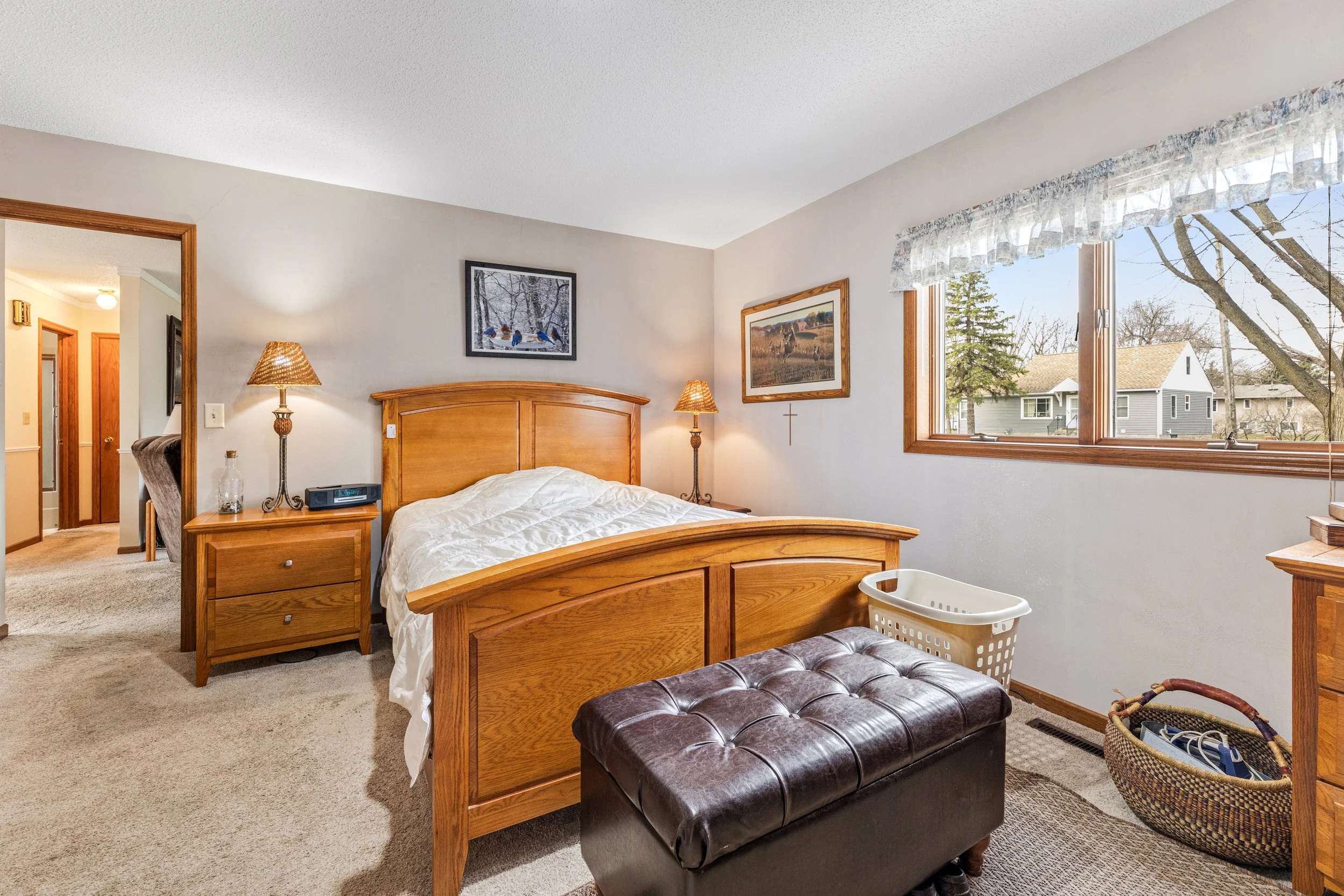 A bedroom with a wooden bed frame, two nightstands with lamps, framed pictures on the wall, a window with a view of houses outside, a brown tufted bench at the foot of the bed, and a laundry basket.