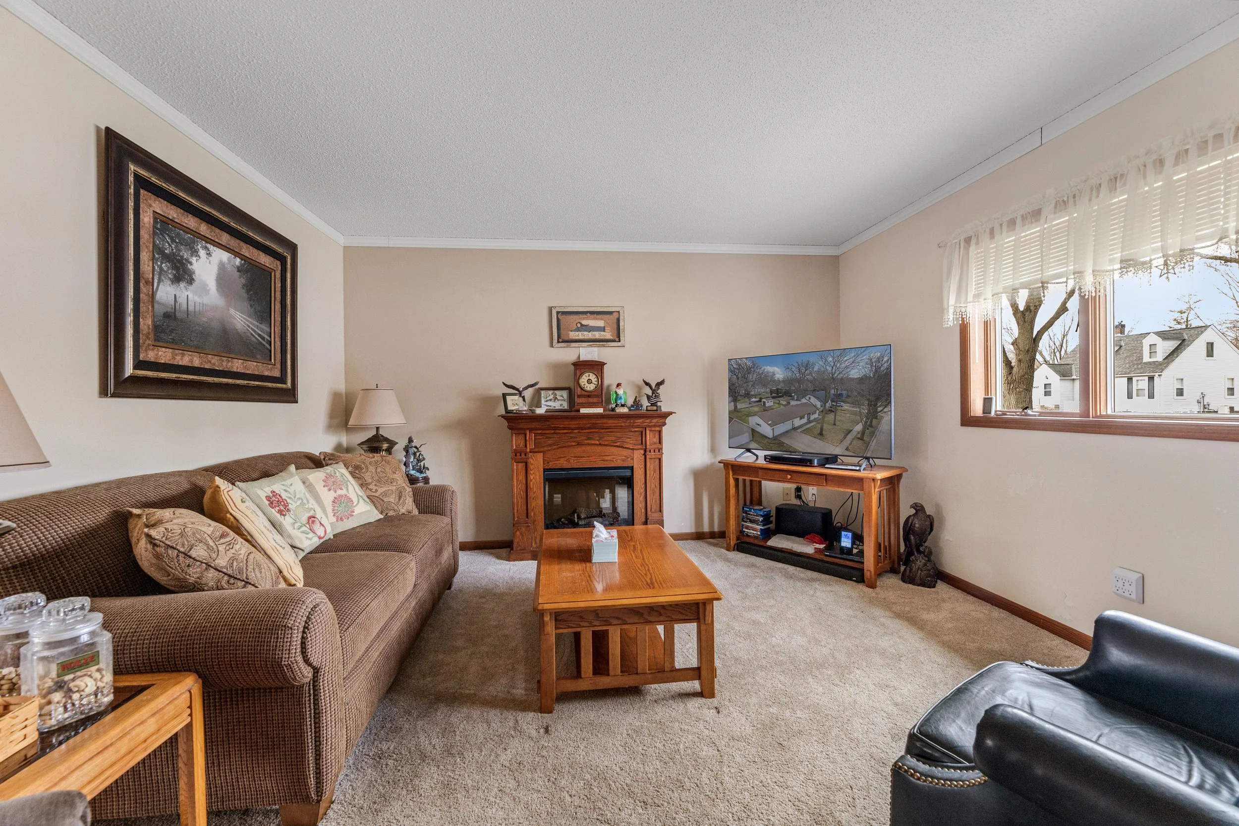Living room with a brown couch, a wooden coffee table, a lamp, family photos, a fireplace, a TV, and a window with blinds showing a view of a house and trees outside.