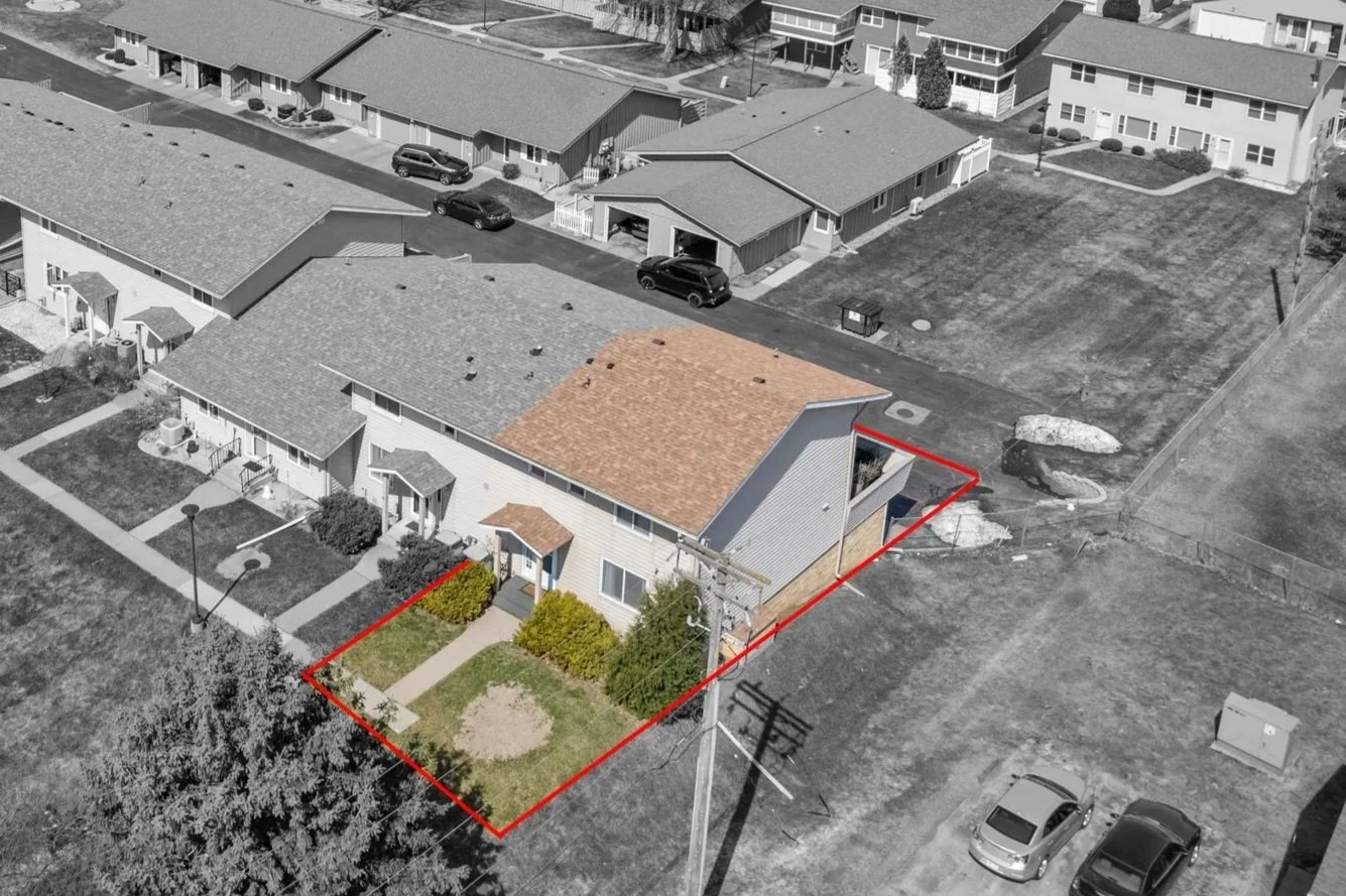 Aerial view of a residential neighborhood with a house outlined in red, showing a small front yard with grass, shrubs, a pathway, and a backyard with a patch of dirt and a small tree. The surrounding area includes other houses, parked cars, and open grassy spaces.