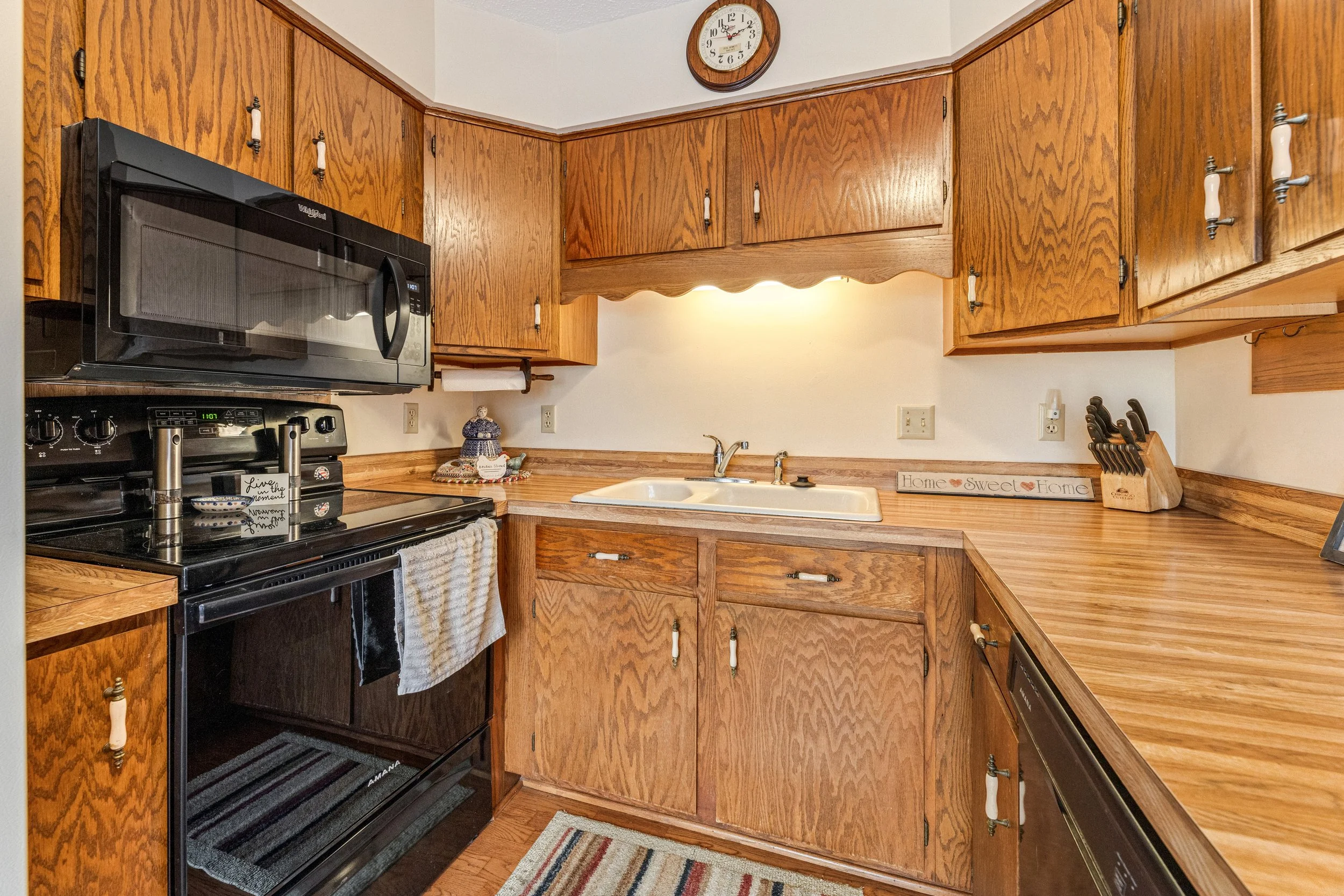 Kitchen with wooden cabinets, black stove, microwave, and a sink. Wall clock showing 1:09. Decorative items and a colorful rug on the floor.