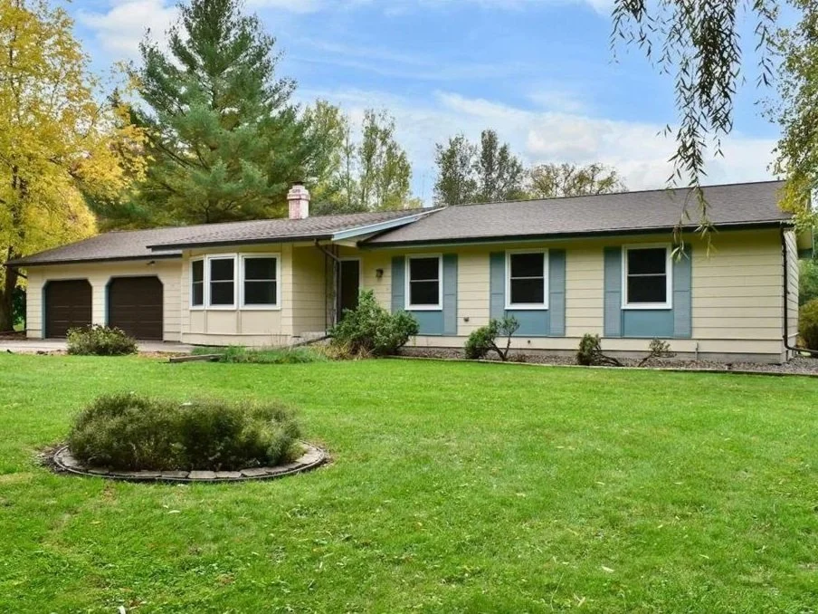 A single-story house with a beige exterior, blue shutters, and a dark gray roof, featuring a two-car garage with dark brown doors, surrounded by a green lawn with bushes and trees in the background under a blue sky.