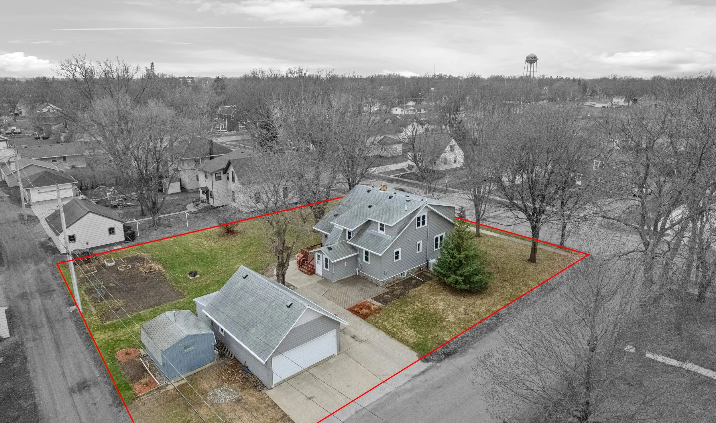 A black and white aerial view of a residential neighborhood with a highlighted house surrounded by a red outline. The house is gray with a gabled roof, a small front porch, and a backyard with a garden plot and a small shed. Bare trees are visible around the property, indicating a winter or early spring season.