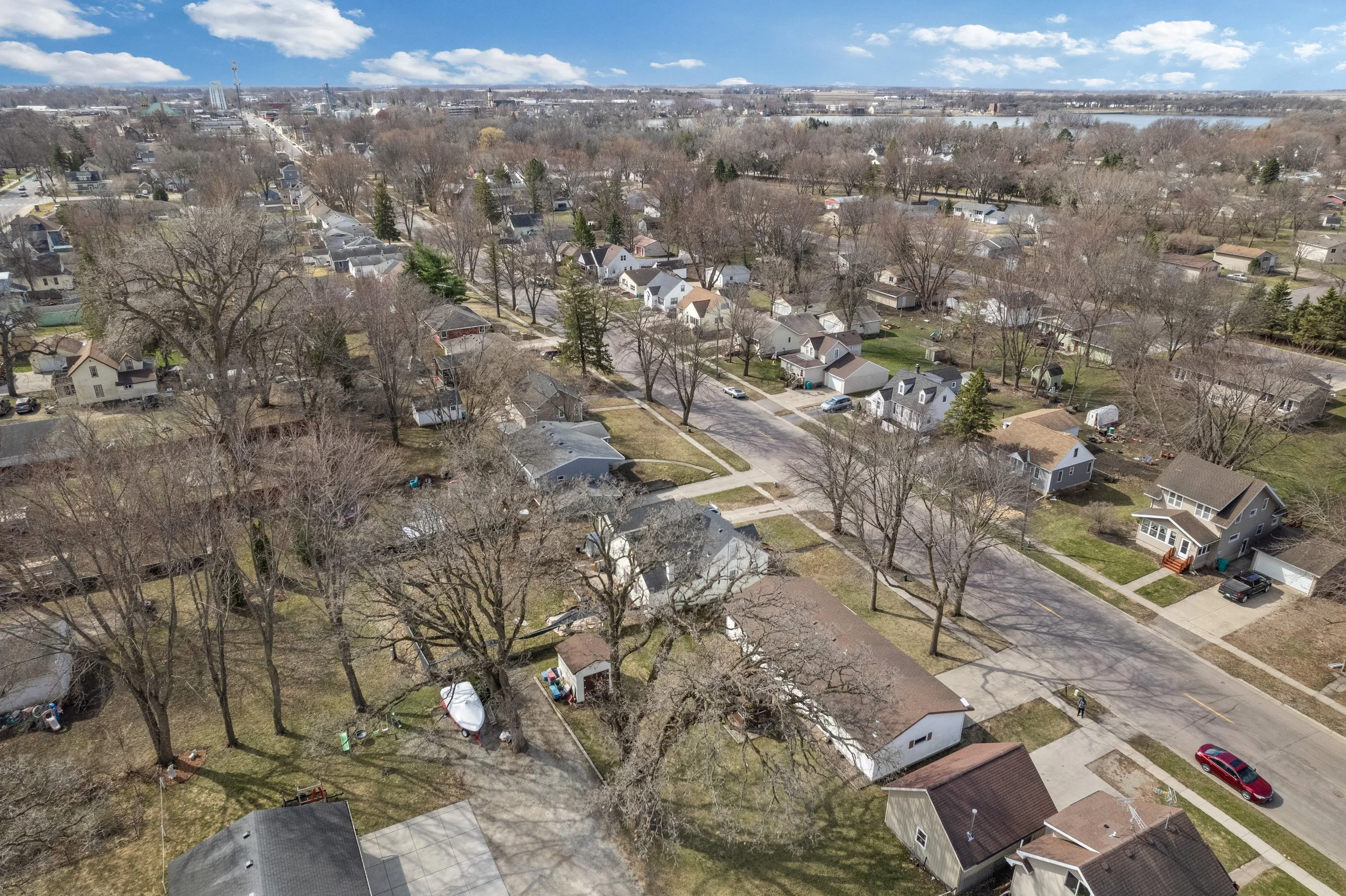 An aerial view of a suburban neighborhood with houses, trees, and streets. The trees are mostly leafless, indicating late fall or winter. There is a river or large body of water in the background under a partly cloudy sky.