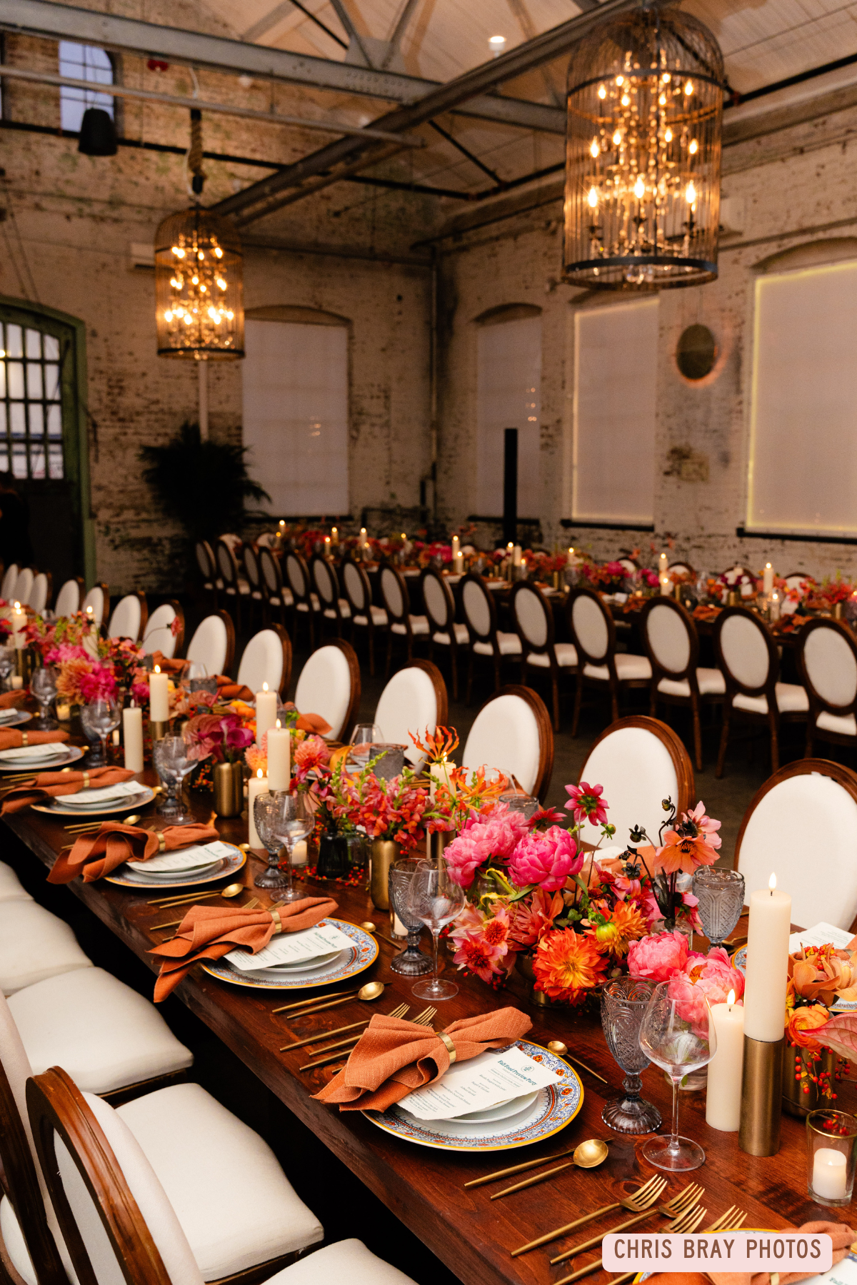 A long banquet table set for a formal event with floral centerpieces, candles, and elegant place settings in a rustic industrial-style venue with large chandeliers.