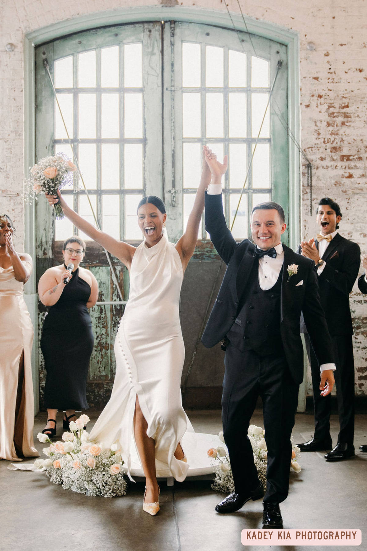 A bride and groom celebrate their wedding with their arms raised, surrounded by friends and family, in an industrial-style venue with large windows and exposed brick walls.