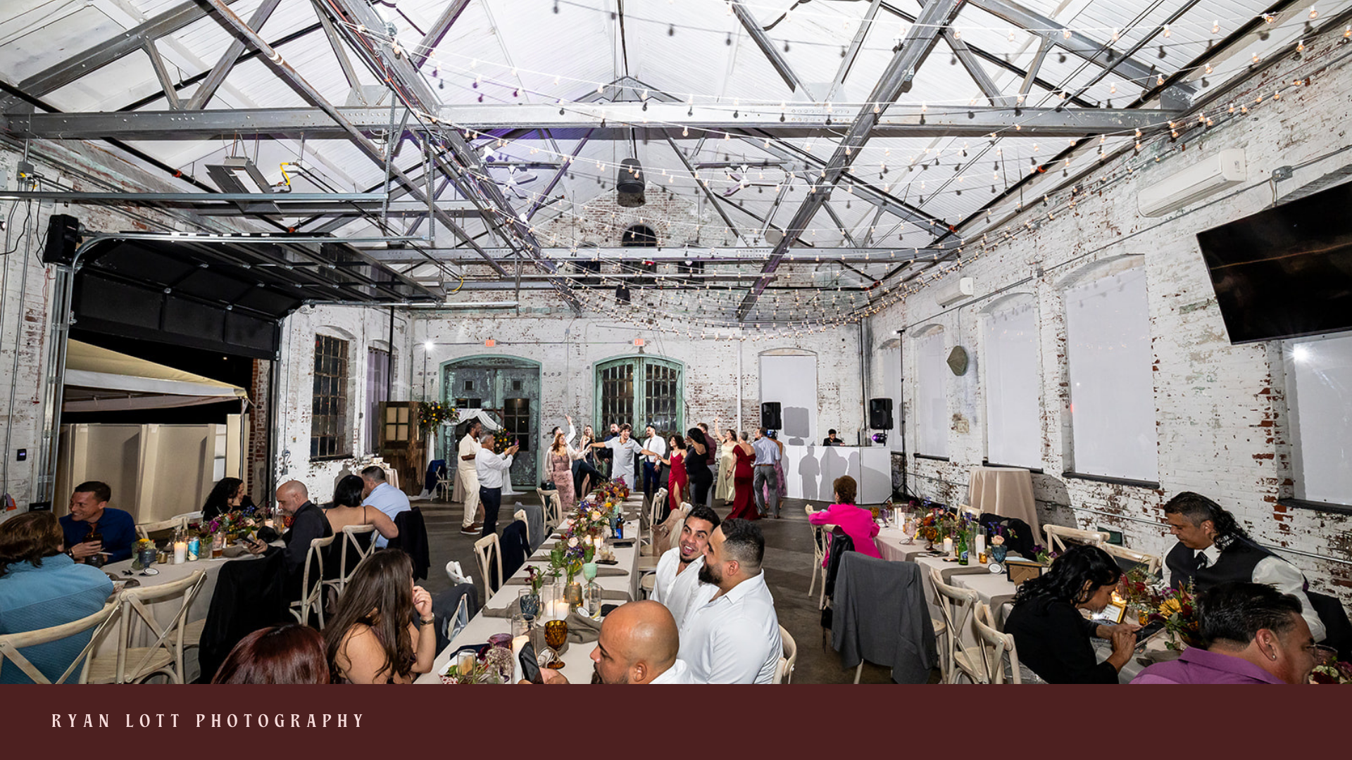 People dancing and celebrating in a rustic, industrial-style event space with exposed brick walls and string lights overhead.