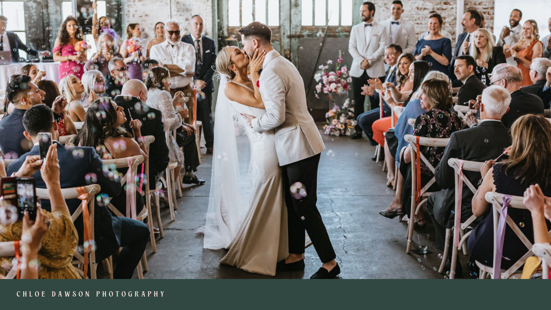 A bride and groom sharing their first dance at a wedding reception, surrounded by seated guests and standing attendants, in a rustic industrial-style venue with exposed brick walls and large windows.