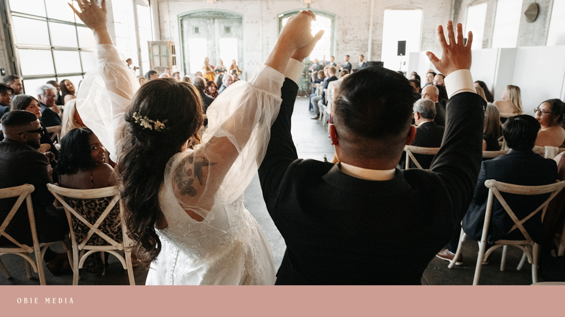 A newly married couple at their wedding reception, dancing with their hands raised in celebration. The bride is wearing a white lace wedding dress with floral tattoos visible on her arm. The groom is dressed in a black suit. They are surrounded by se