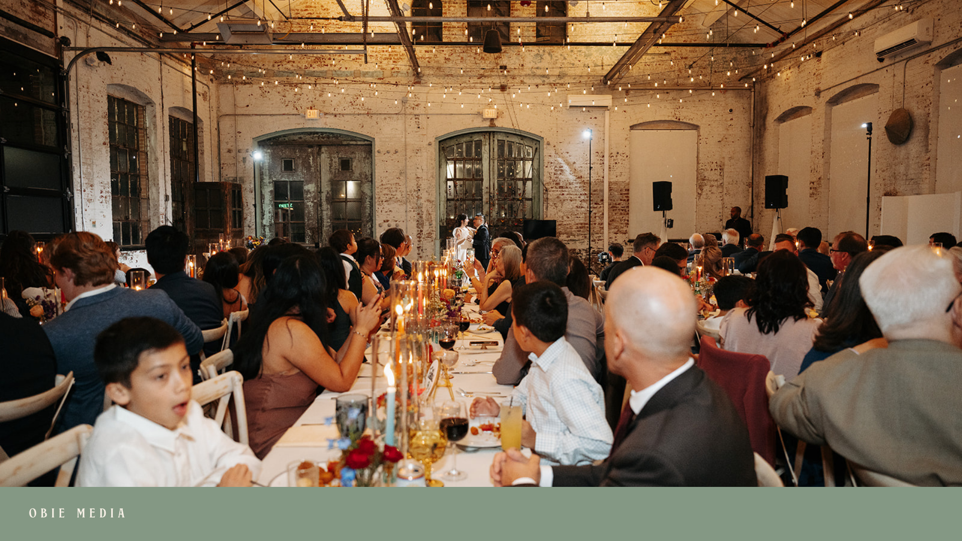 The image shows a large indoor gathering, possibly a wedding reception, with many guests seated at long tables decorated with candles, flowers, and drinks. The venue has exposed brick walls, large windows, and string lights on the ceiling, creating a