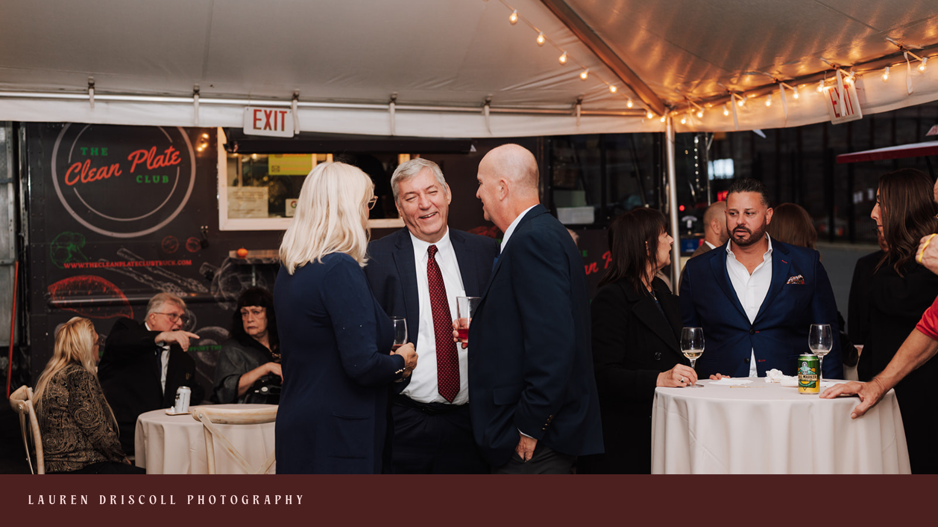 People gathered at an outdoor event under a tent, engaging in conversation. The scene features a mix of men and women in business attire, some holding wine glasses. A food truck named 'The Clean Plate Club' is visible in the background, with string l