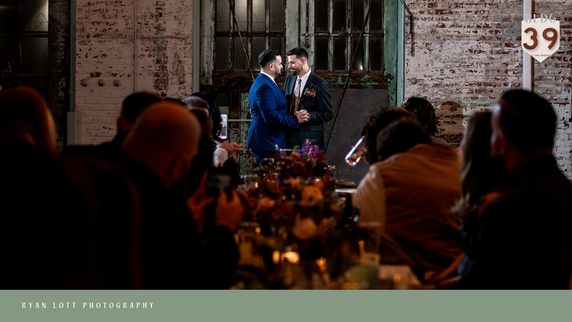 Two men in formal attire holding hands and smiling at each other during a wedding ceremony, with guests seated around watching in an industrial-style venue with exposed brick walls.