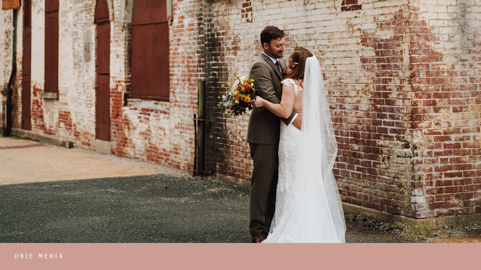 A bride and groom stand facing each other against a brick wall, with the bride holding a colorful bouquet of flowers. The bride is wearing a white lace wedding dress and veil, and the groom is dressed in a brown suit. They are smiling and looking int