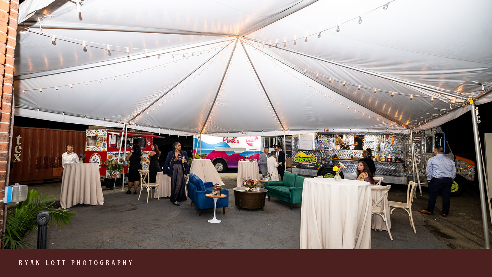 Outdoor or tented event setup with string lights hanging from a large canopy. There are round cocktail tables with white tablecloths, a green and a blue sofa, and a small round table with a flower arrangement. Several people are standing, conversing,