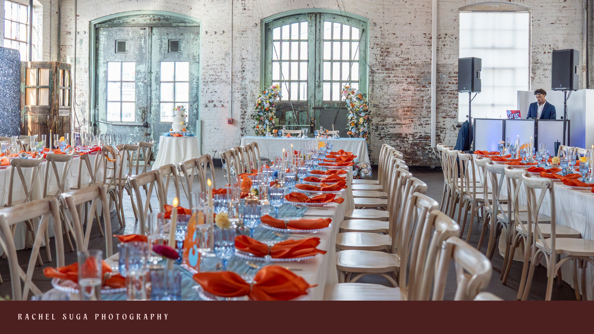 Wedding reception setup in an industrial space with long tables decorated with white tablecloths, orange napkins, and candles; floral arch and wedding cake near rustic green doors; DJ booth and speakers in background.