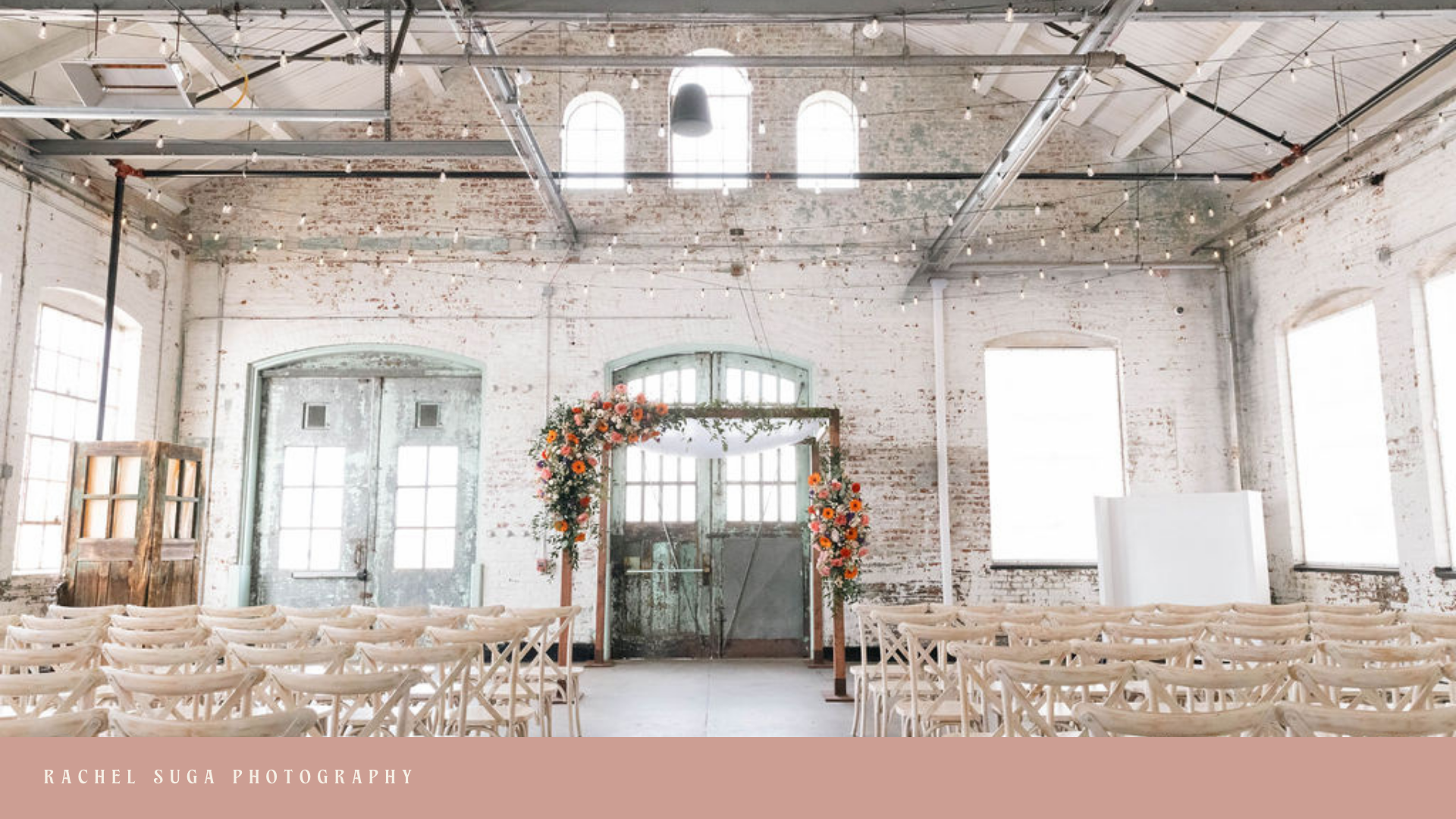 Indoor wedding ceremony setup in a rustic, industrial-style venue with white brick walls, large windows, wooden chairs arranged facing a floral arch, and string lights hanging from the ceiling.