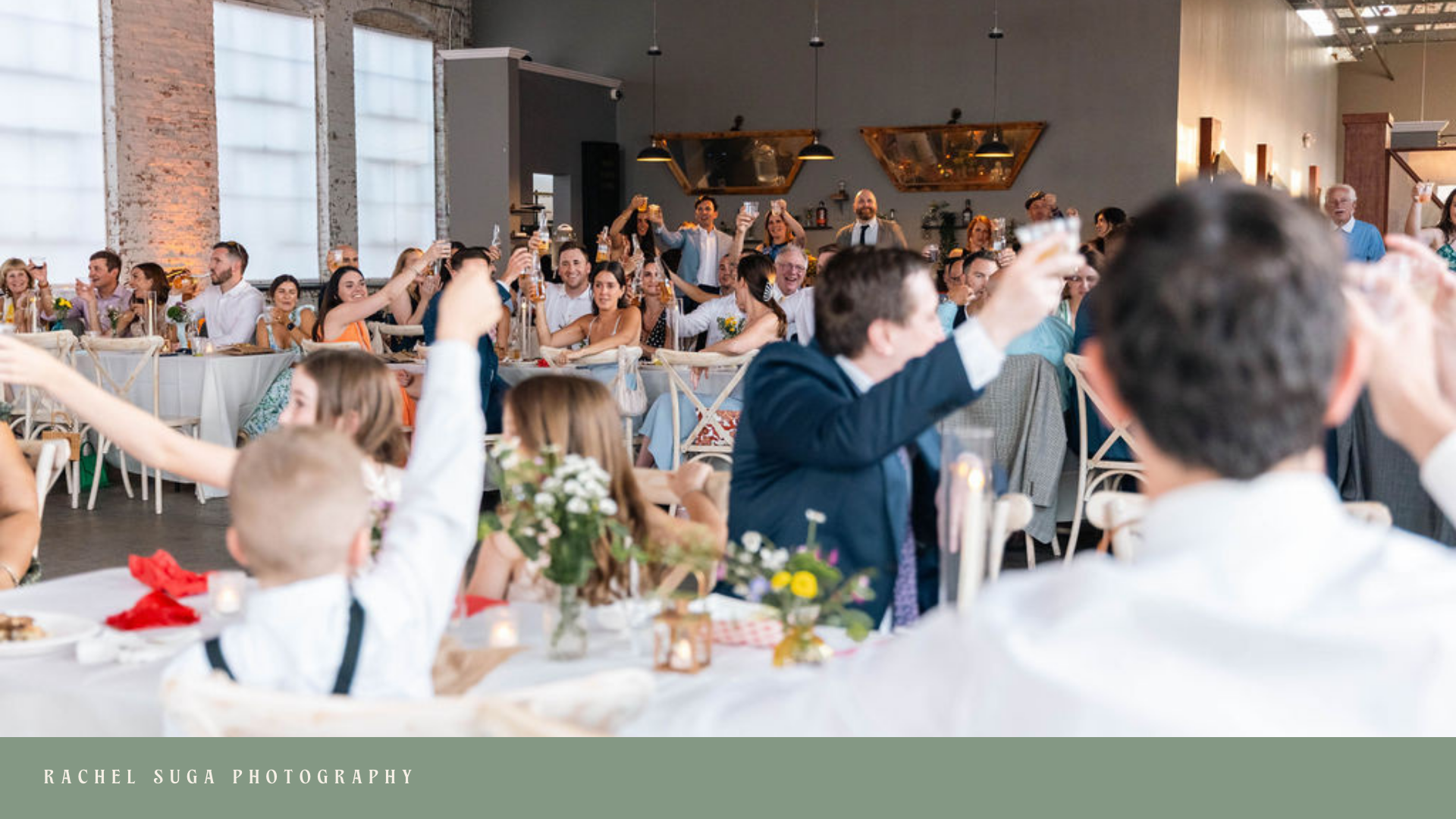 Celebration at a wedding reception with guests raising glasses in toast, seated at decorated tables in a spacious, industrial-style venue.