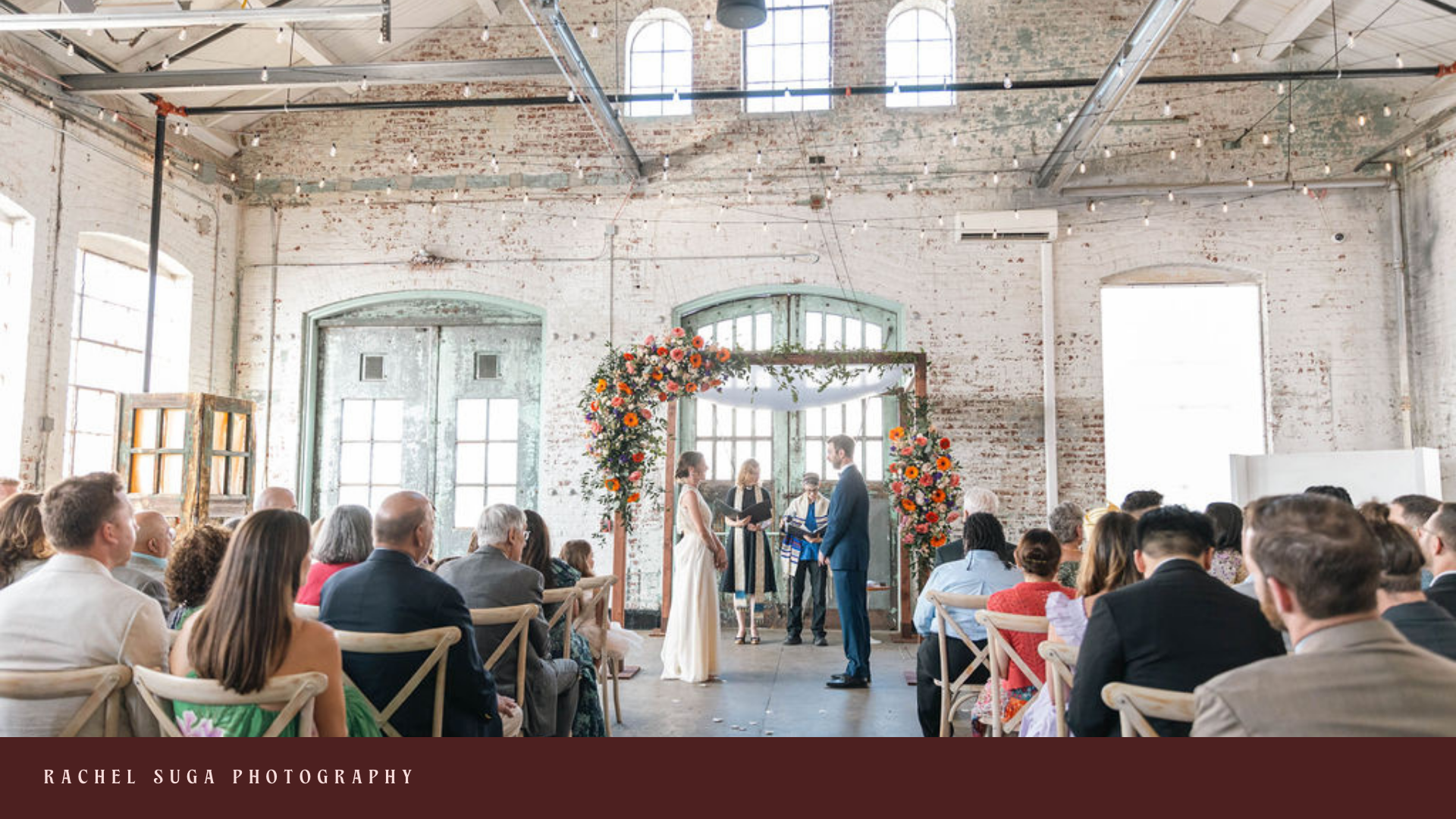 A wedding ceremony taking place in an industrial-style venue with exposed brick walls and large windows. The bride and groom are standing under a floral arch, facing each other, with an officiant and another person present. Guests are seated and watc