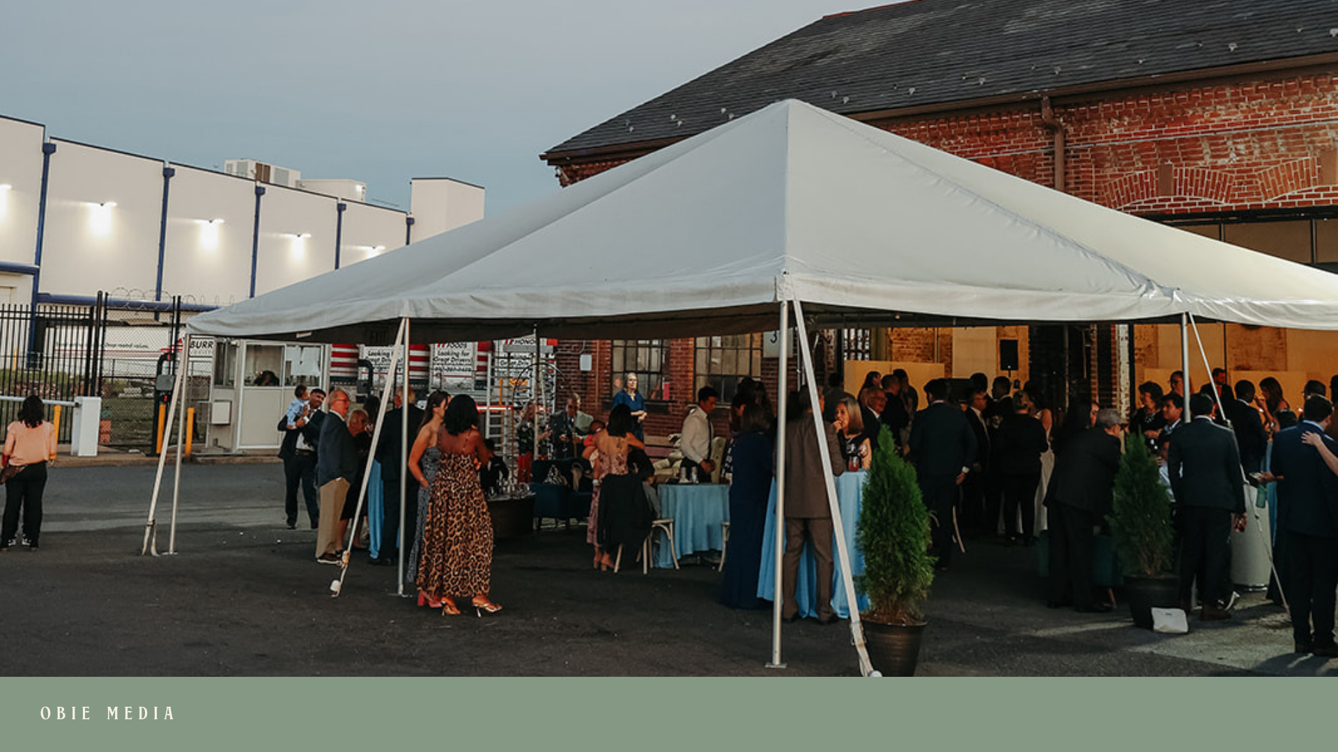 People gathered at an outdoor event under a large white canopy tent, with tables and speakers, in front of a brick building during evening.