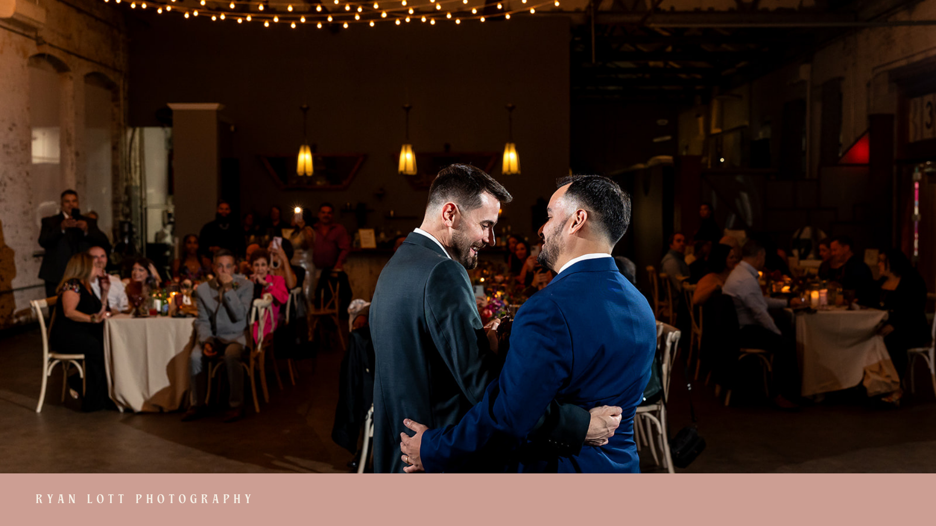 Two men in suits sharing a dance at a wedding reception, surrounded by seated guests in a warmly lit banquet hall with hanging pendant lights.