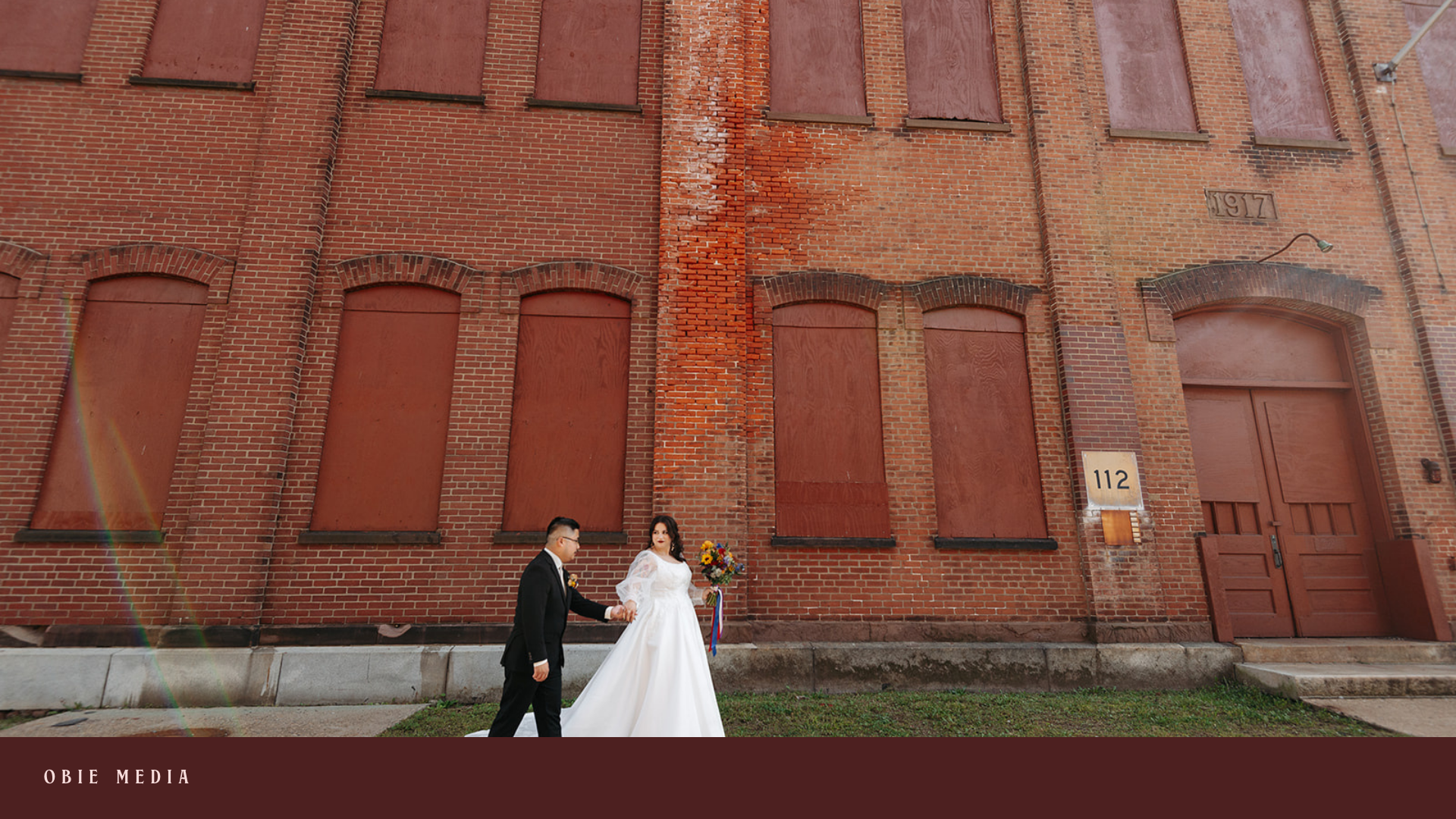 A bride and groom holding hands outside a red brick building with boarded-up windows. The bride is in a white wedding dress holding a bouquet, and the groom is in a black suit.