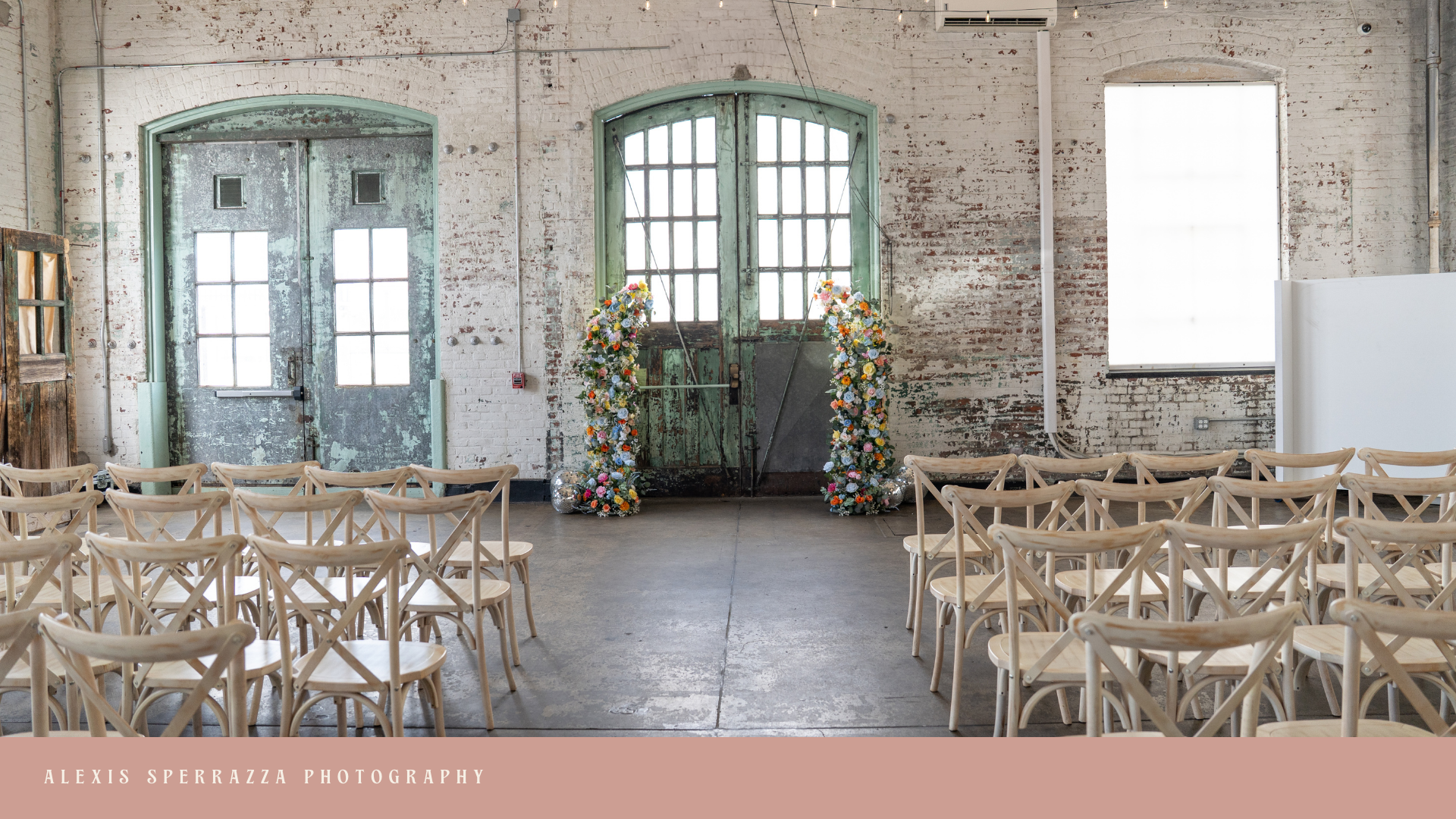 An industrial-style wedding ceremony setup with rows of light wooden chairs facing a vintage, distressed green metal door decorated with two floral archways made of various colorful flowers. The room has exposed brick walls, large windows, and a conc