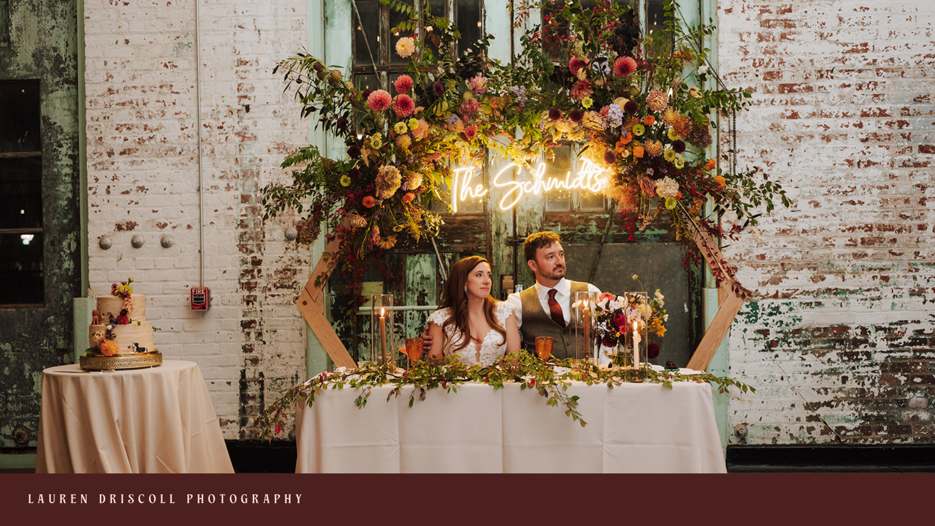 A wedding reception with a bride and groom sitting at a decorated table. Behind them is a floral arch with a neon sign that reads 'The Schultzs'. There is a wedding cake on a separate table to the left and the background features a rustic brick wall.