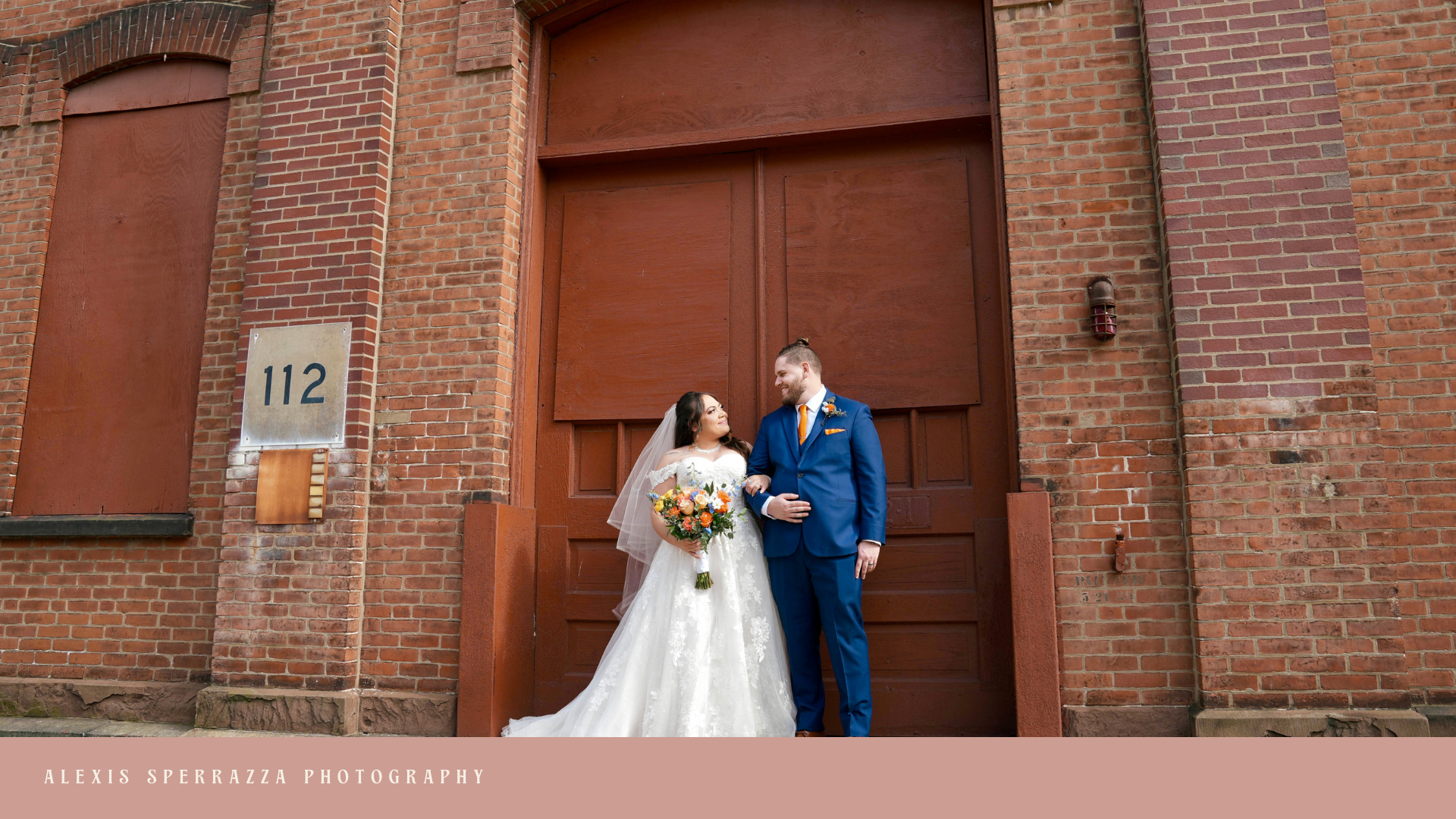 Bride and groom standing arm in arm in front of a brick building with a large wooden door, smiling at each other, she holding a bouquet of flowers, he wearing a blue suit with an orange tie.