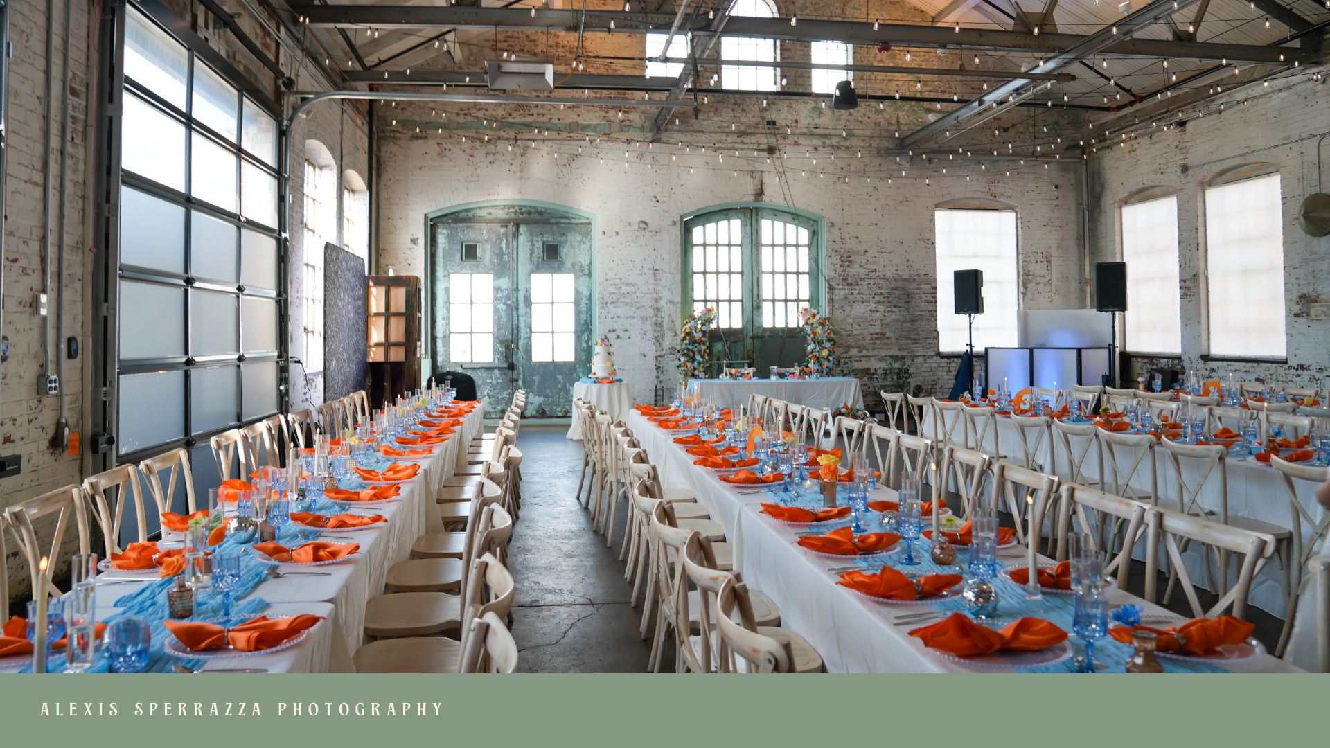 Event space decorated with long banquet tables set with orange napkins, clear blue glasses, and small floral arrangements, inside an industrial-style building with exposed brick walls and large windows.
