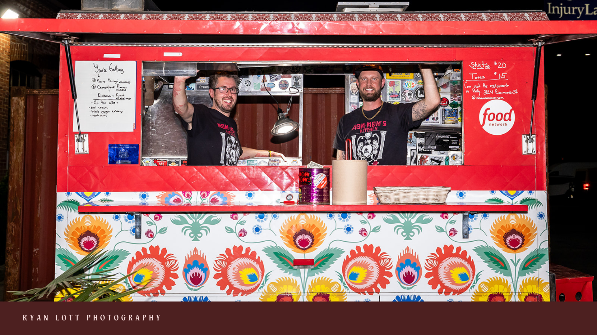 Two smiling men inside a colorful food truck with a floral pattern, wearing black t-shirts that say "Mom-Mom's Kitchen."