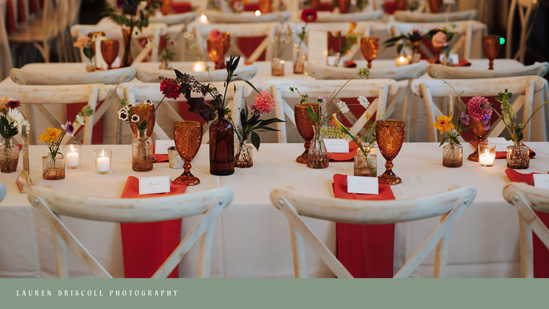 Elegant event table decorated with amber glassware, small candles, and colorful flowers in vases, set with white tablecloths and red cloth napkins, with rustic white chairs, in a warm, ambient setting.