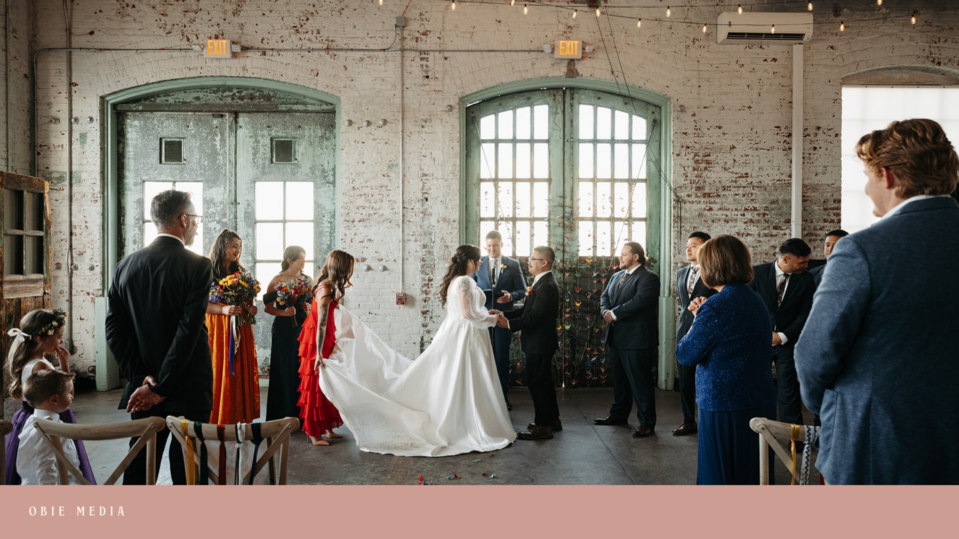 A wedding ceremony inside an industrial-style building with brick walls and large industrial windows. The bride and groom are holding hands at the altar, surrounded by friends and family, with the bride wearing a white wedding gown and the groom in a