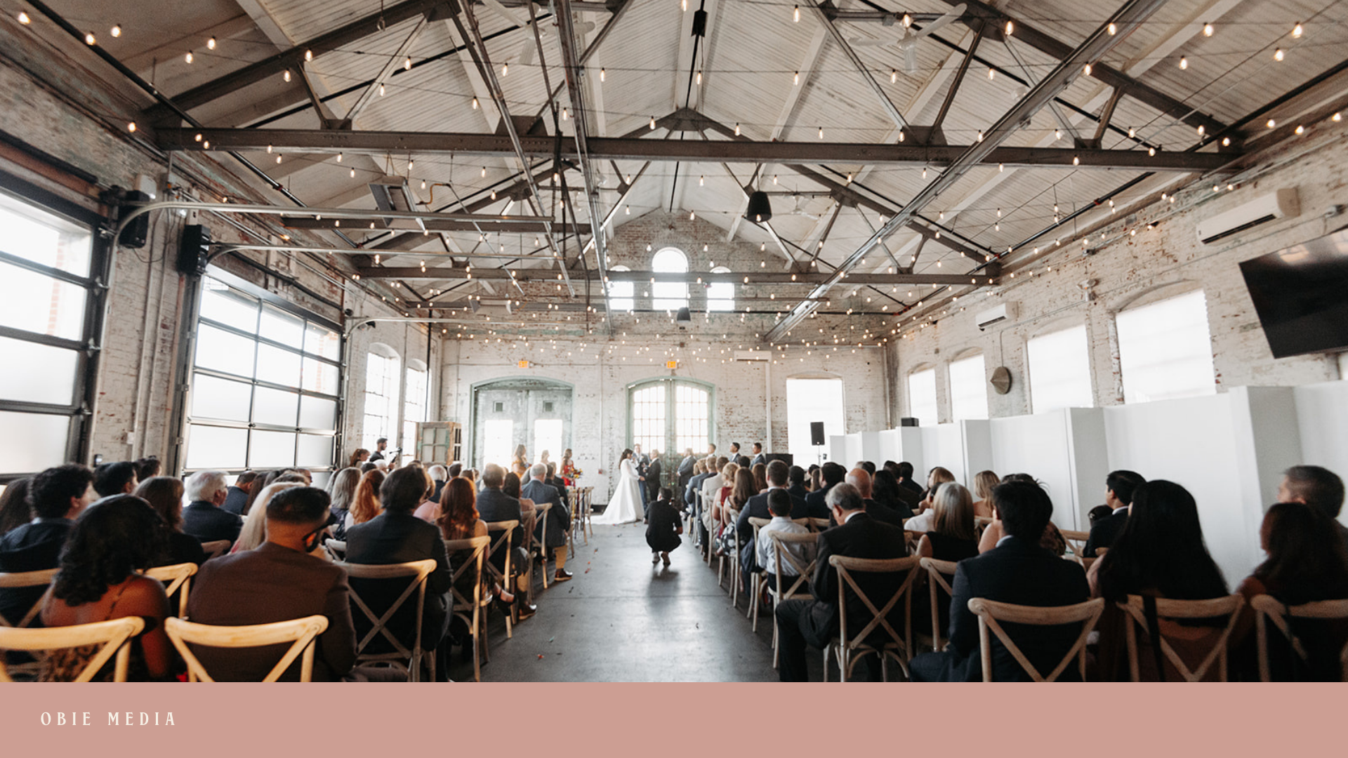 Wedding ceremony inside a spacious industrial-style venue with high ceilings, string lights, and large windows. The bride and groom are standing at the altar in front of the officiant, with guests seated on wooden chairs facing them.