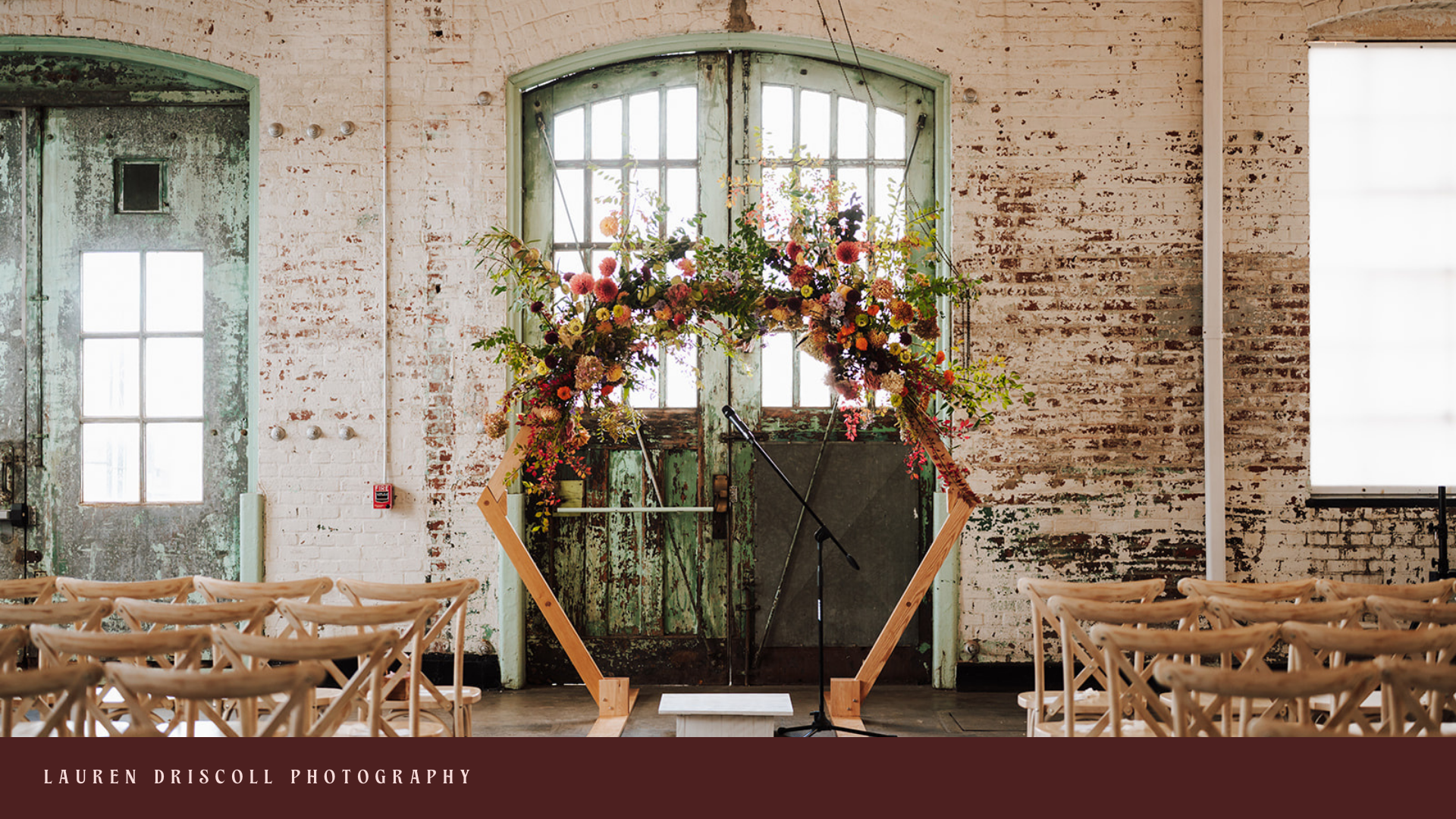 Decorated arch with flowers set up on a stage in front of rustic green double doors and large windows in an industrial-style venue, with wooden chairs arranged for an event.