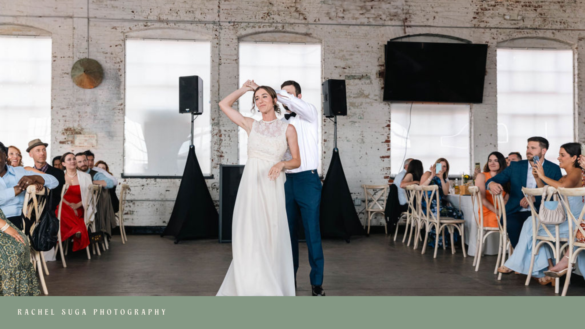 A bride and groom sharing their first dance at a wedding reception in a rustic venue with white brick walls and large windows, surrounded by seated guests watching and taking photos.
