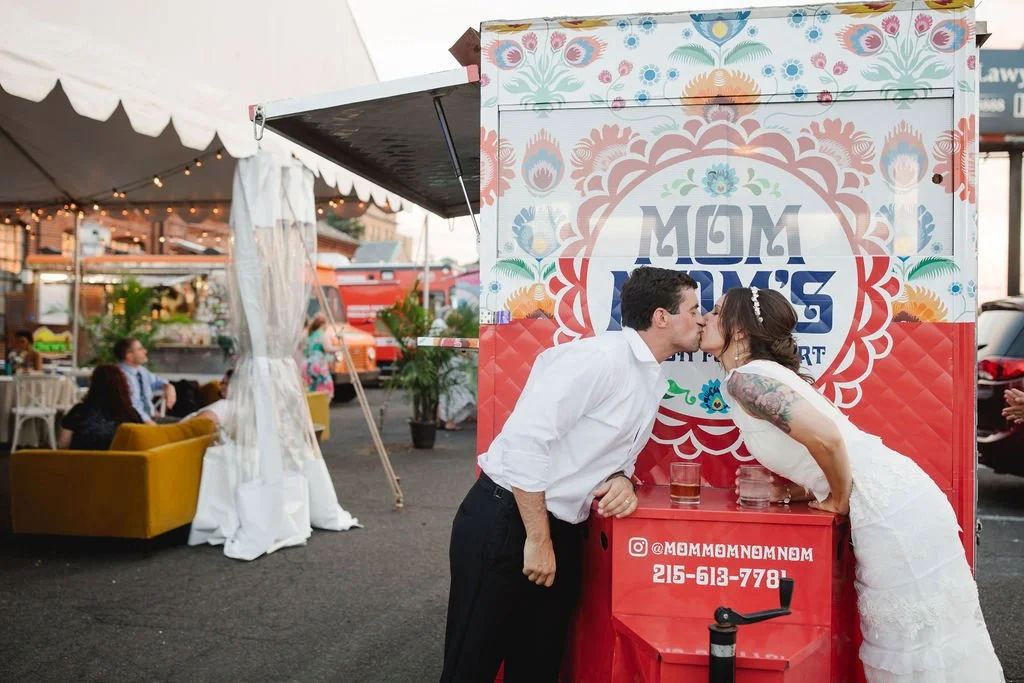 A couple kissing in front of a colorful food truck at an outdoor event, with people sitting on yellow couches and festive string lights overhead.