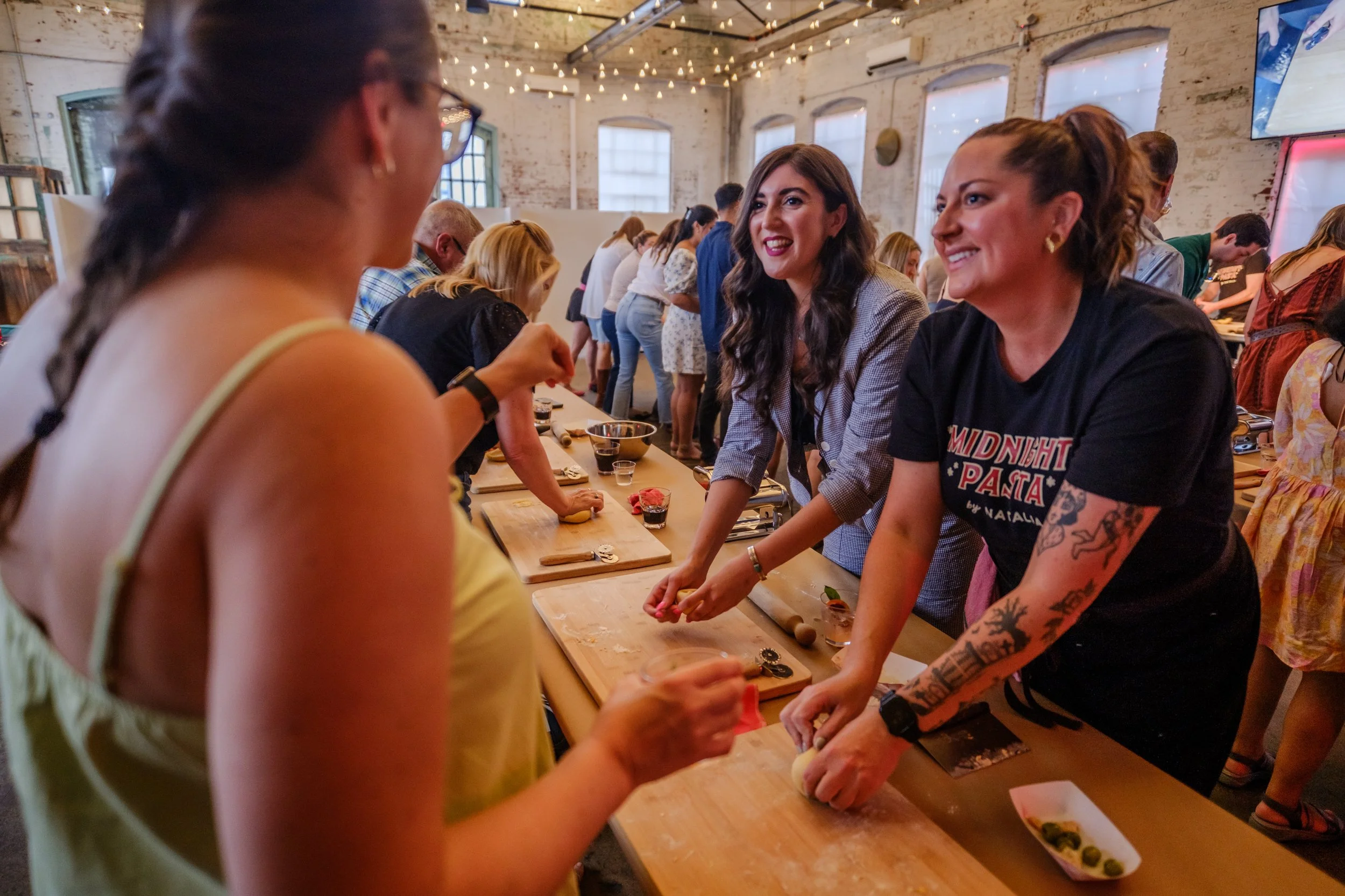 People participating in a cooking class or demonstration, rolling dough at a long wooden table in a rustic, industrial-style venue with string lights and large windows.