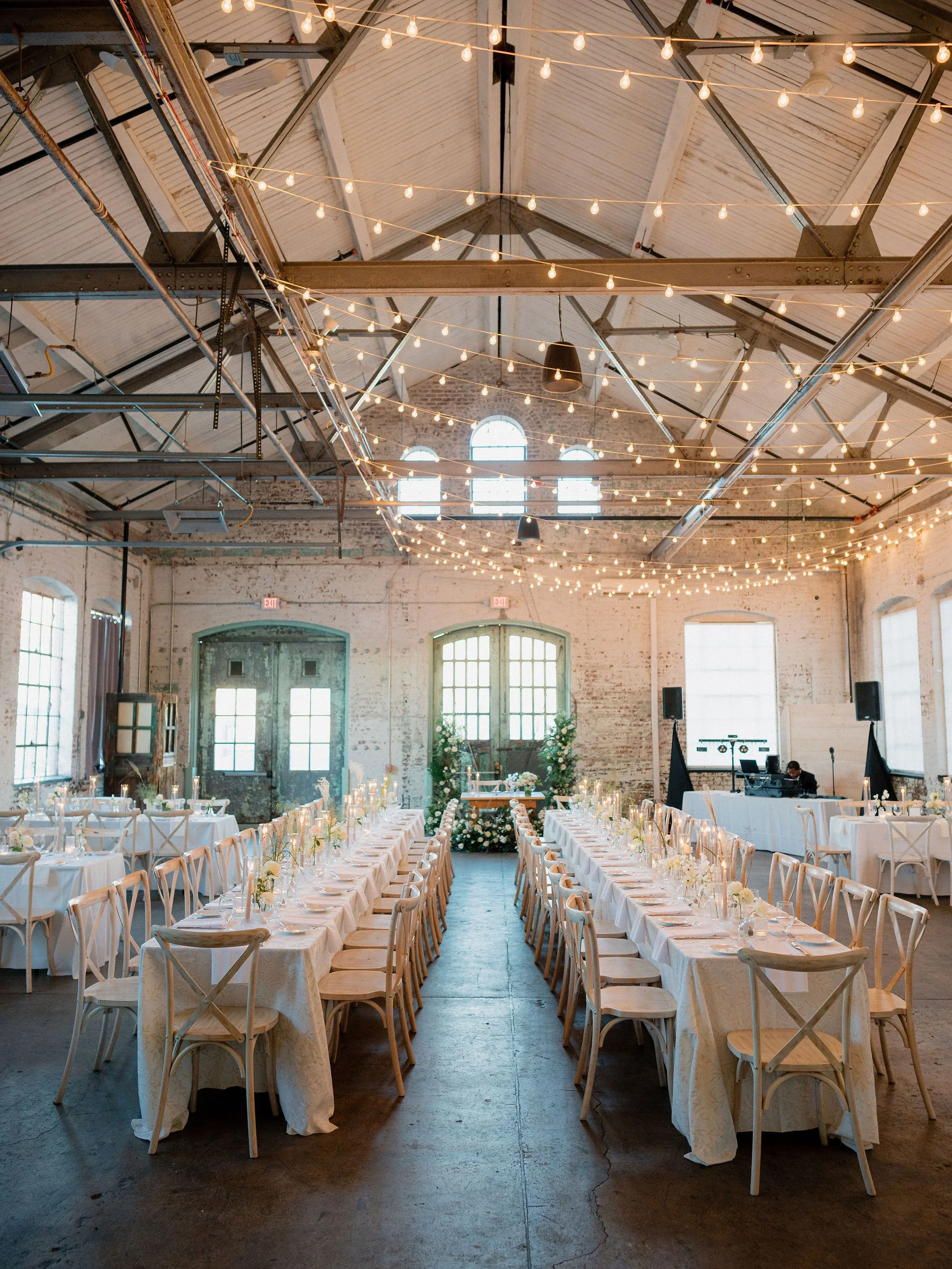 A long banquet table set for a formal event with floral centerpieces, candles, and elegant place settings in a rustic industrial-style venue with large chandeliers.