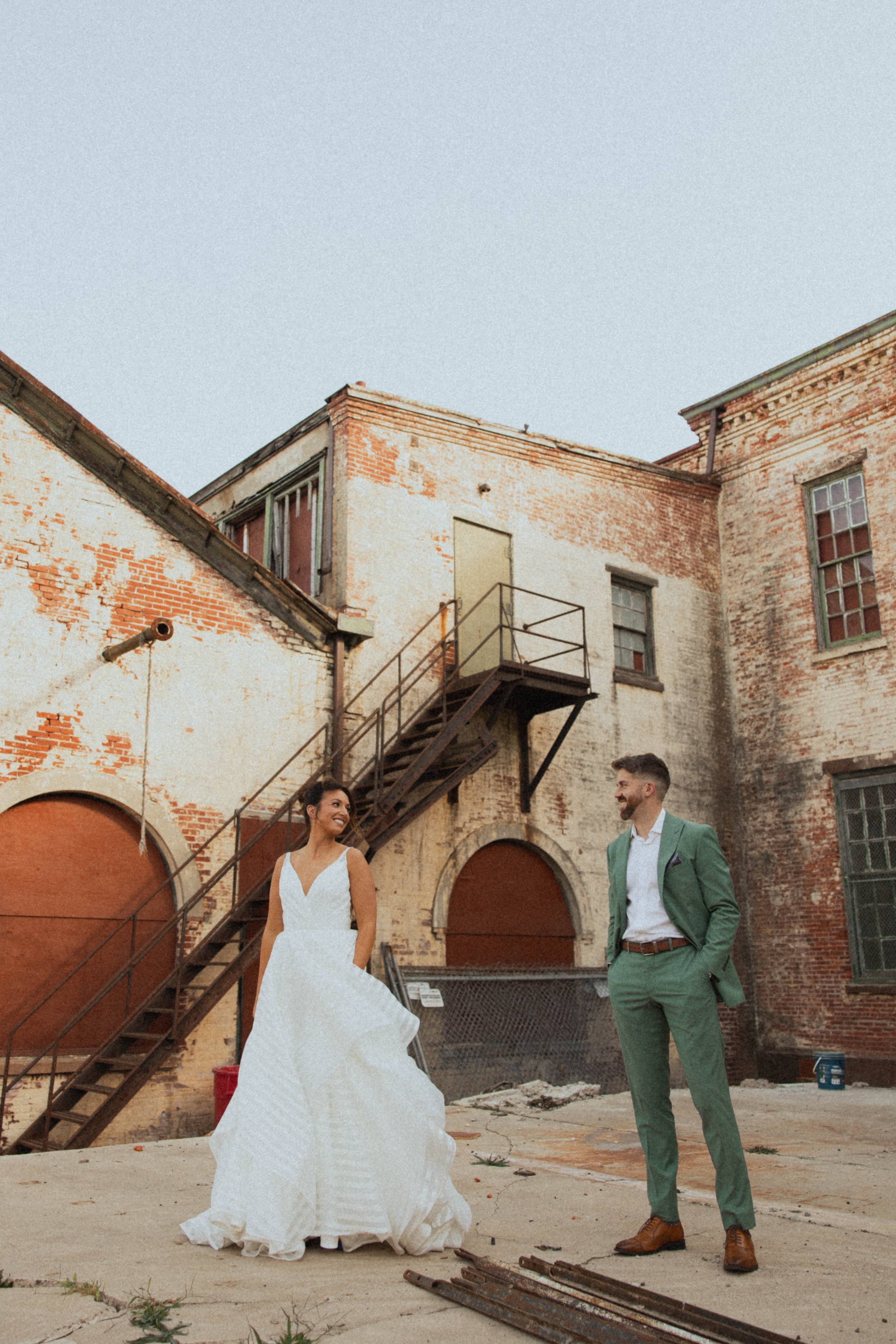 A bride and groom celebrate their wedding with their arms raised, surrounded by friends and family, in an industrial-style venue with large windows and exposed brick walls.