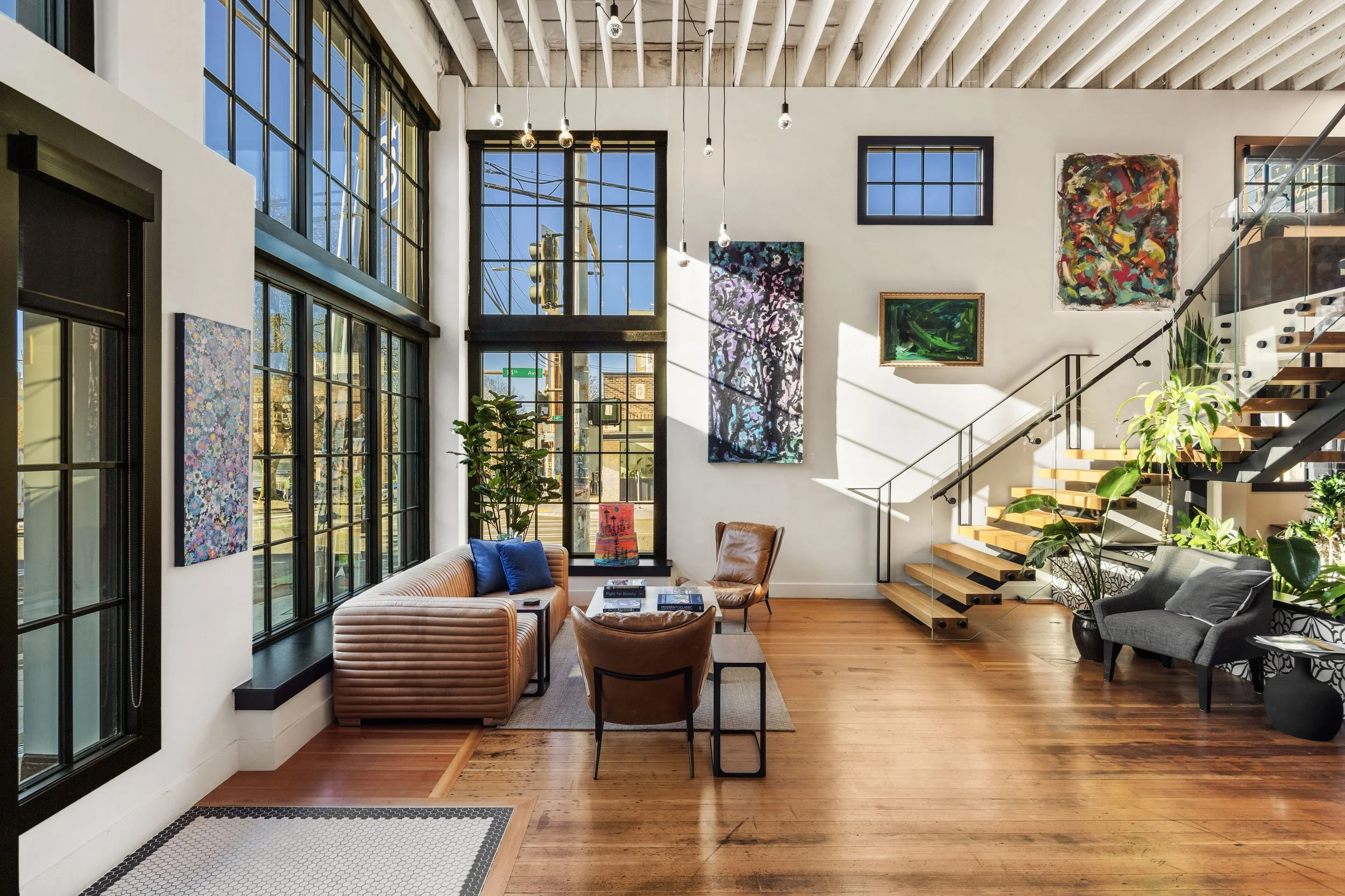 Bright living room with large windows, wooden flooring, and modern furniture, including a sofa, chairs, and artwork on the walls.