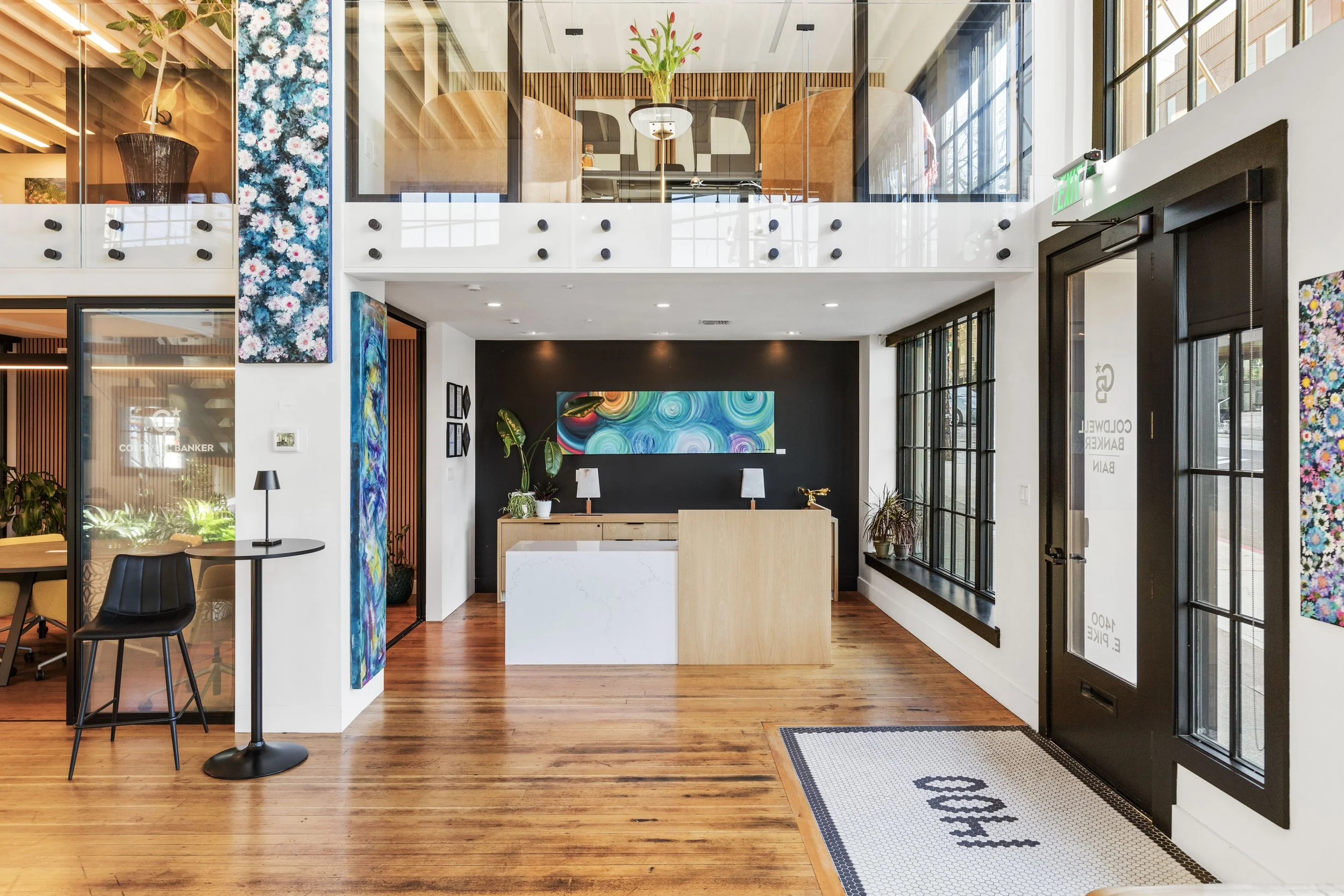 Modern and stylish hotel lobby with wooden floors, artwork, plants, a reception desk, large windows, and a small seating area near the entrance.