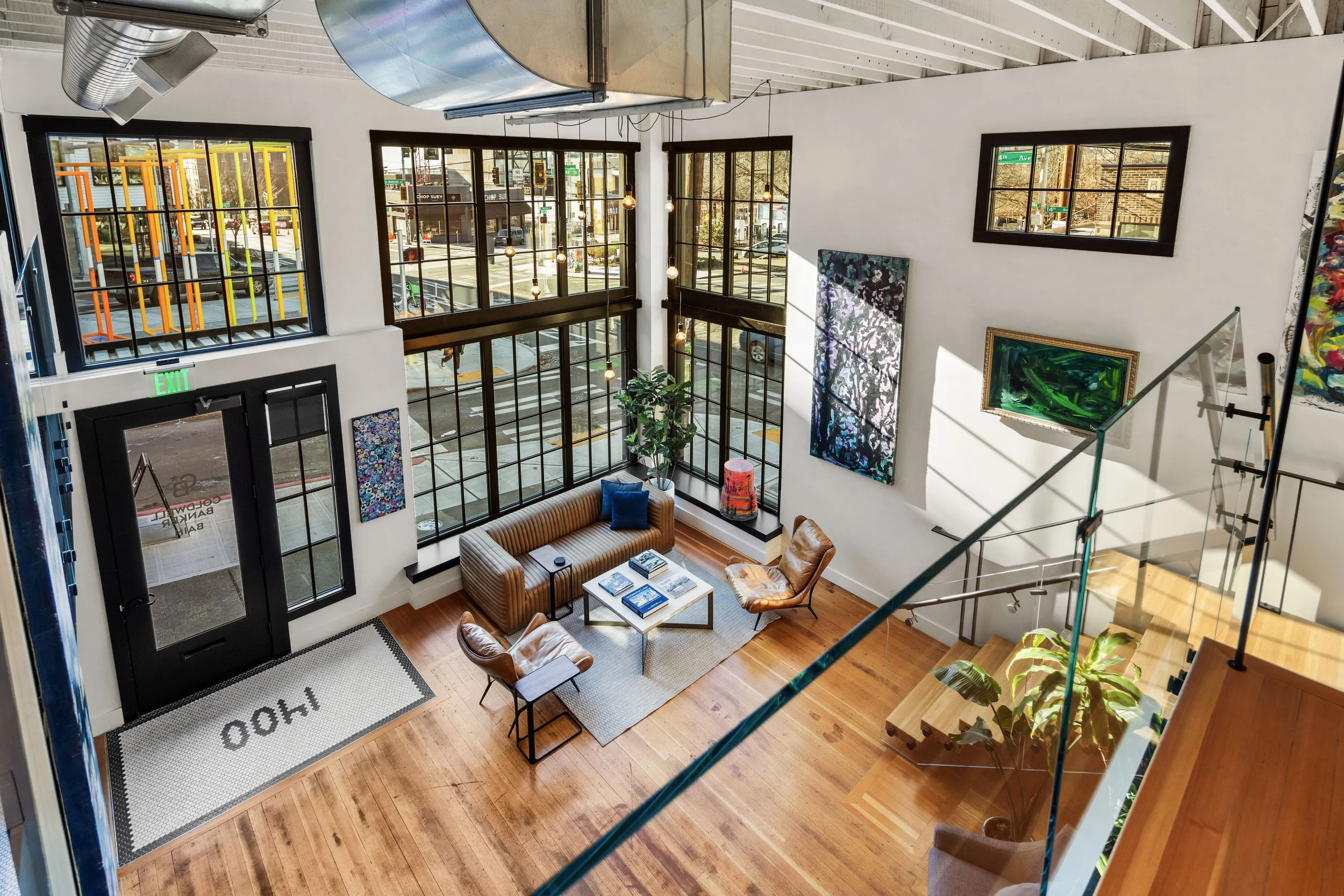 Interior of a modern art gallery with large windows, wooden flooring, a brown striped sofa, leather chairs, and colorful artwork on white walls.