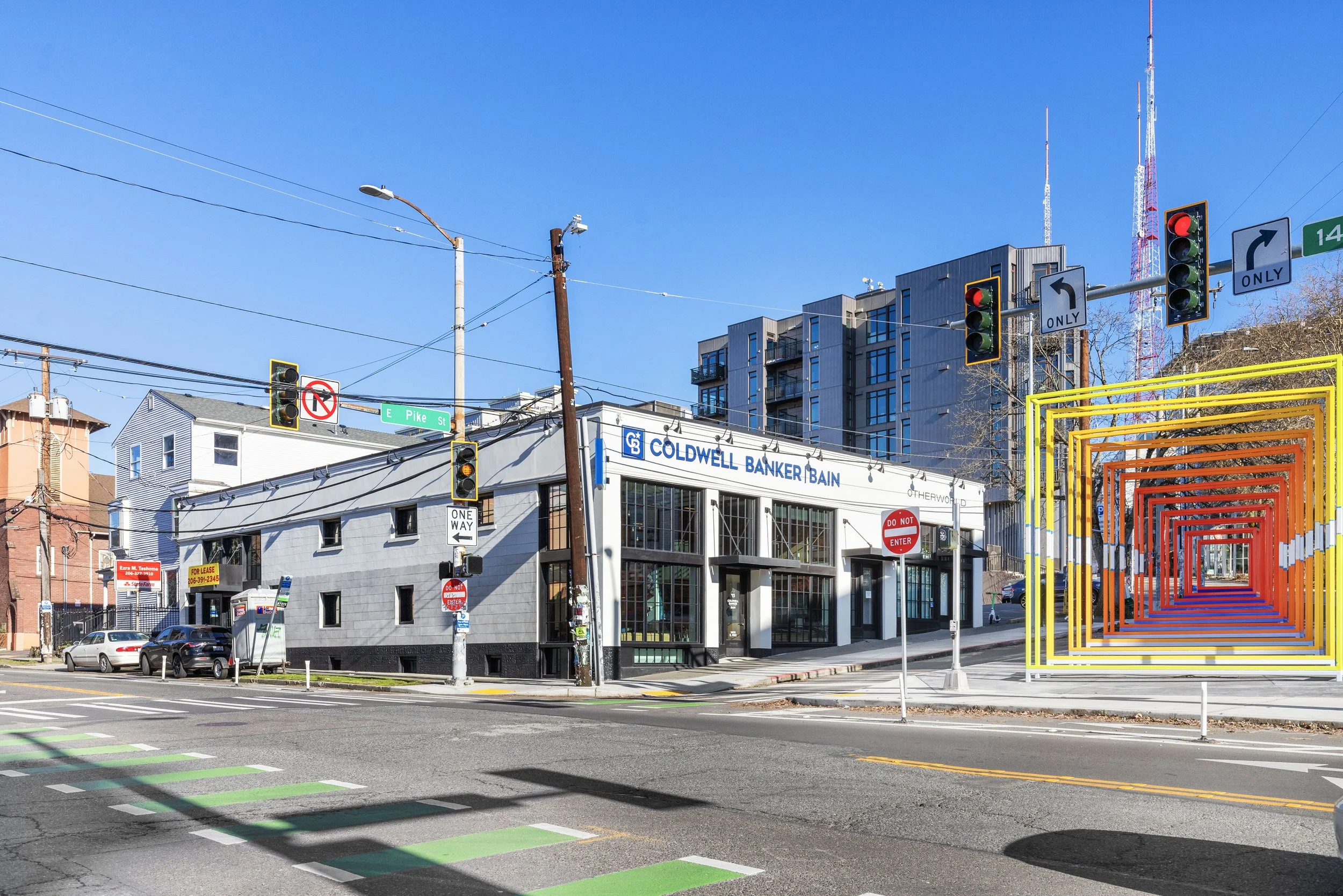 City street corner with traffic lights, signs, a building labeled Coldwell Banker Bain, and colorful geometric arch structures on the sidewalk.