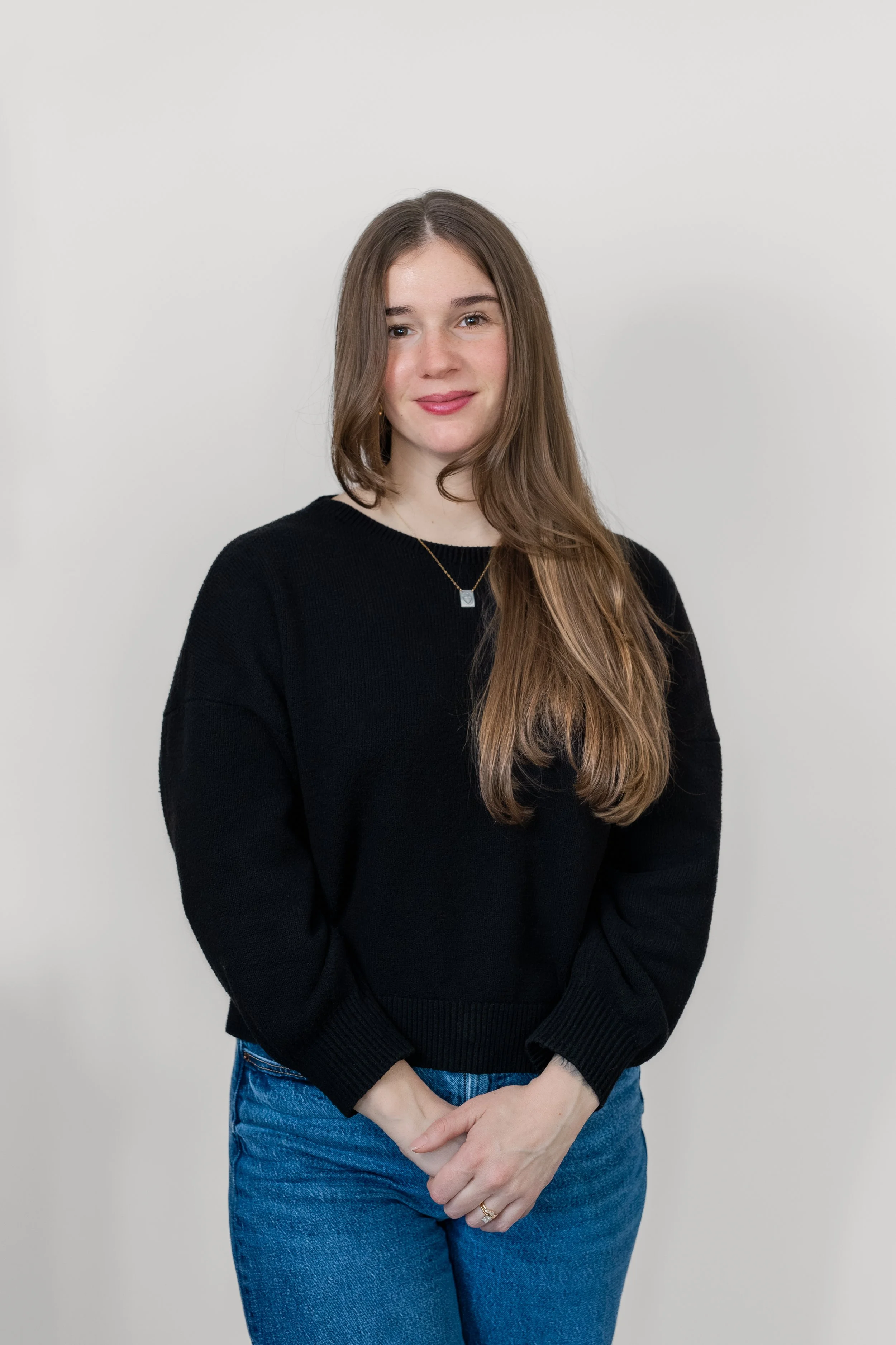 A young woman with long brown hair, wearing a black sweater and blue jeans, standing against a plain light gray background.