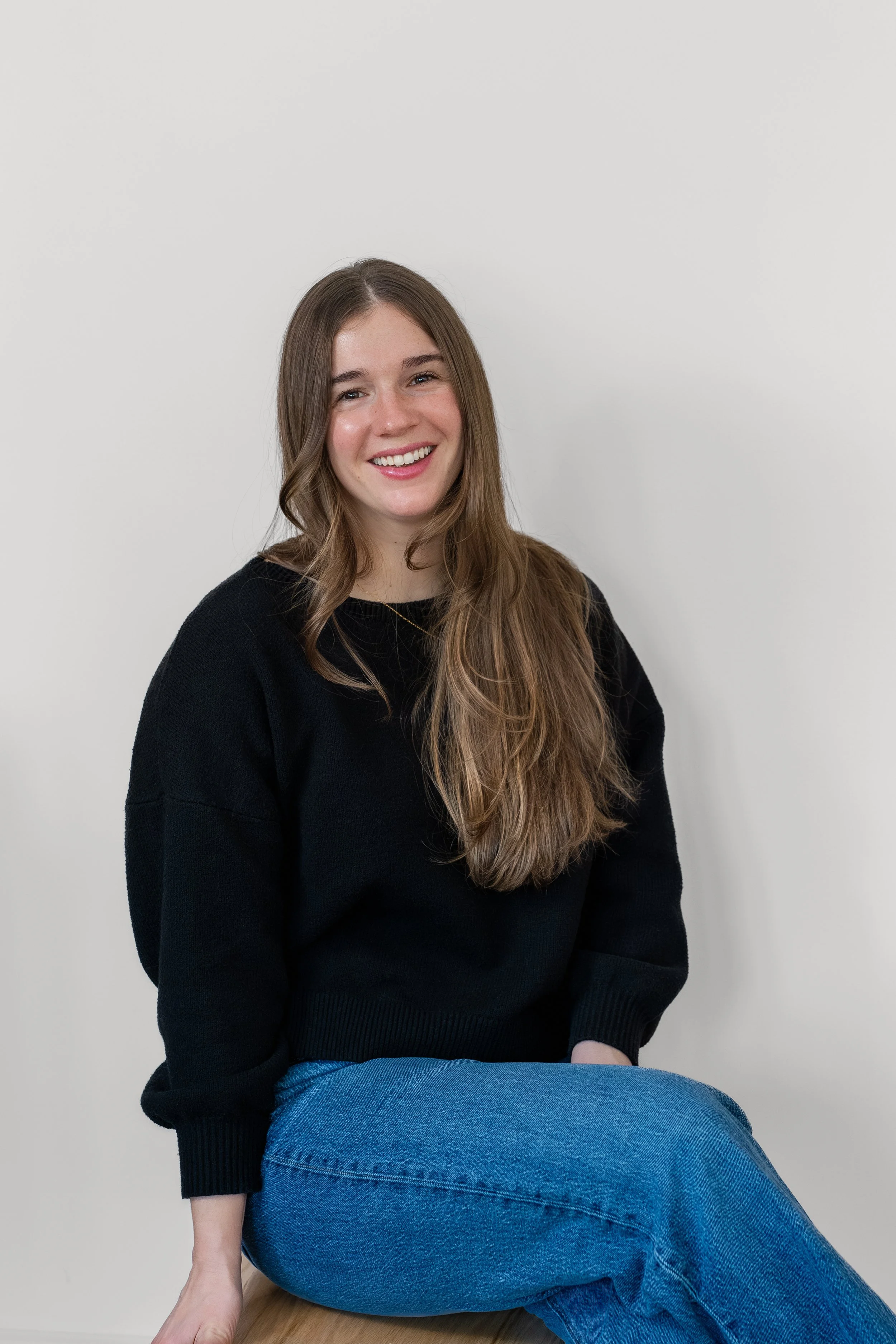 A young woman with long brown hair, wearing a black sweater and blue jeans, sitting on a wooden stool against a plain white wall, smiling at the camera.