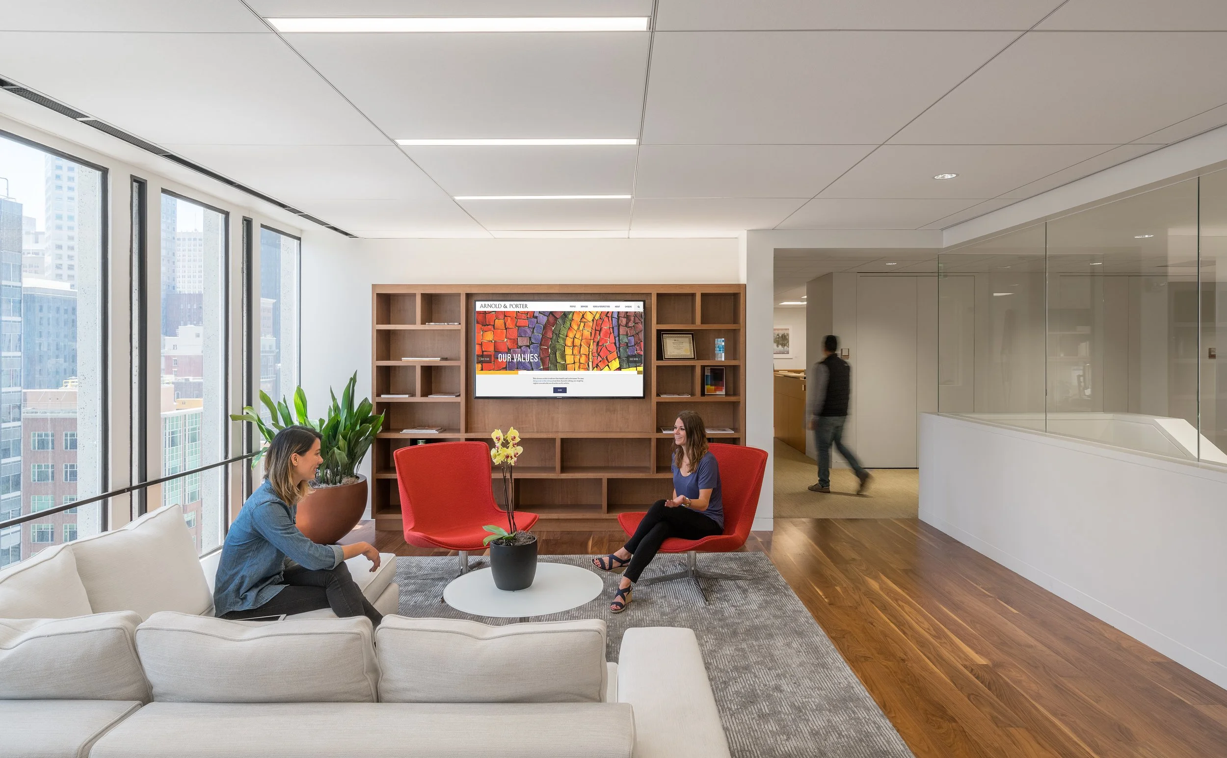 Modern office lounge area with large windows, white sofa, red armchairs, a round coffee table with a plant, and a large wooden bookshelf with a screen displaying colorful artwork and text in the background. Two women are sitting and talking, one walking in the background.