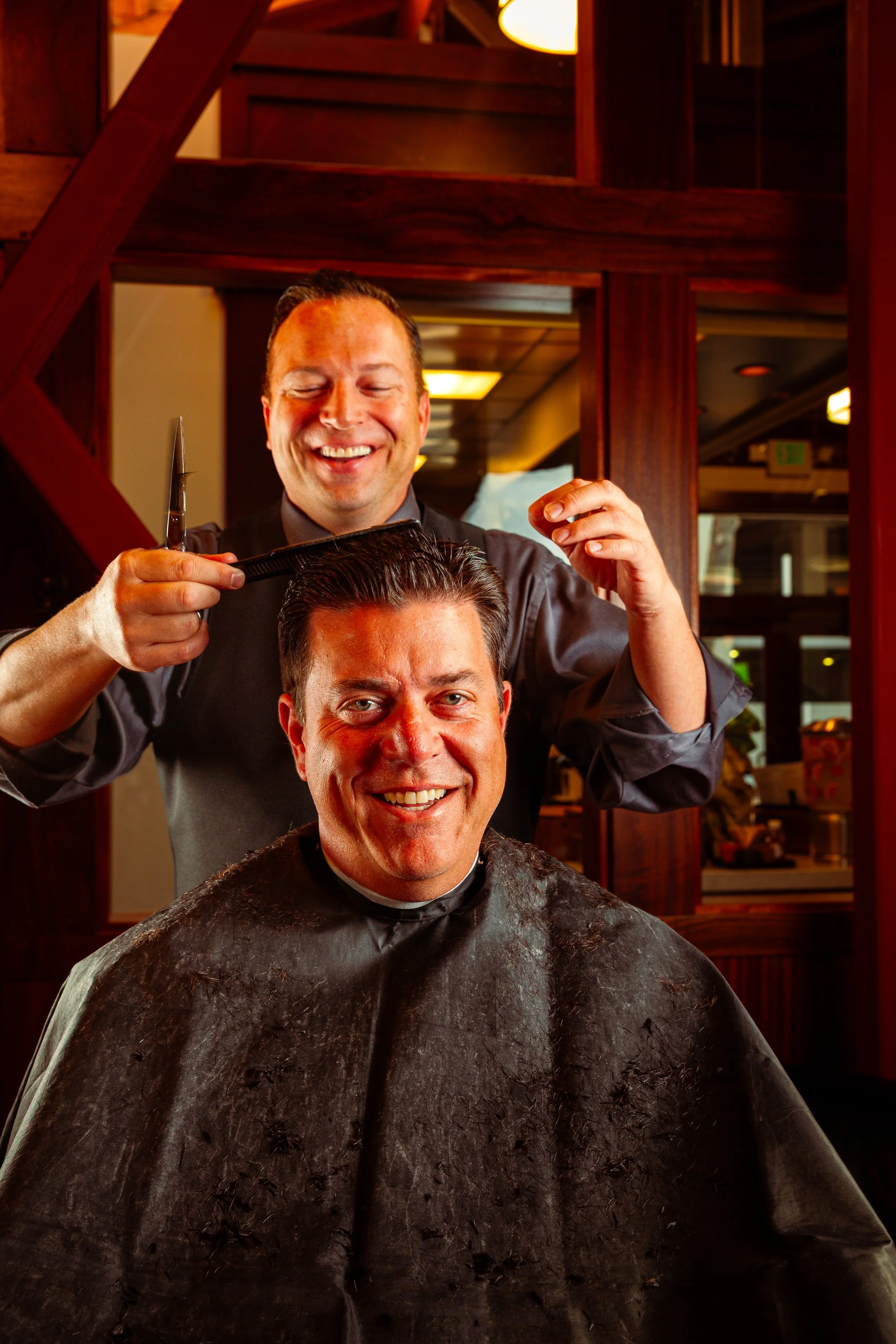 A man getting a haircut at a hair salon, smiling as the barber cuts his hair, with mirrors and warm lighting in the background.