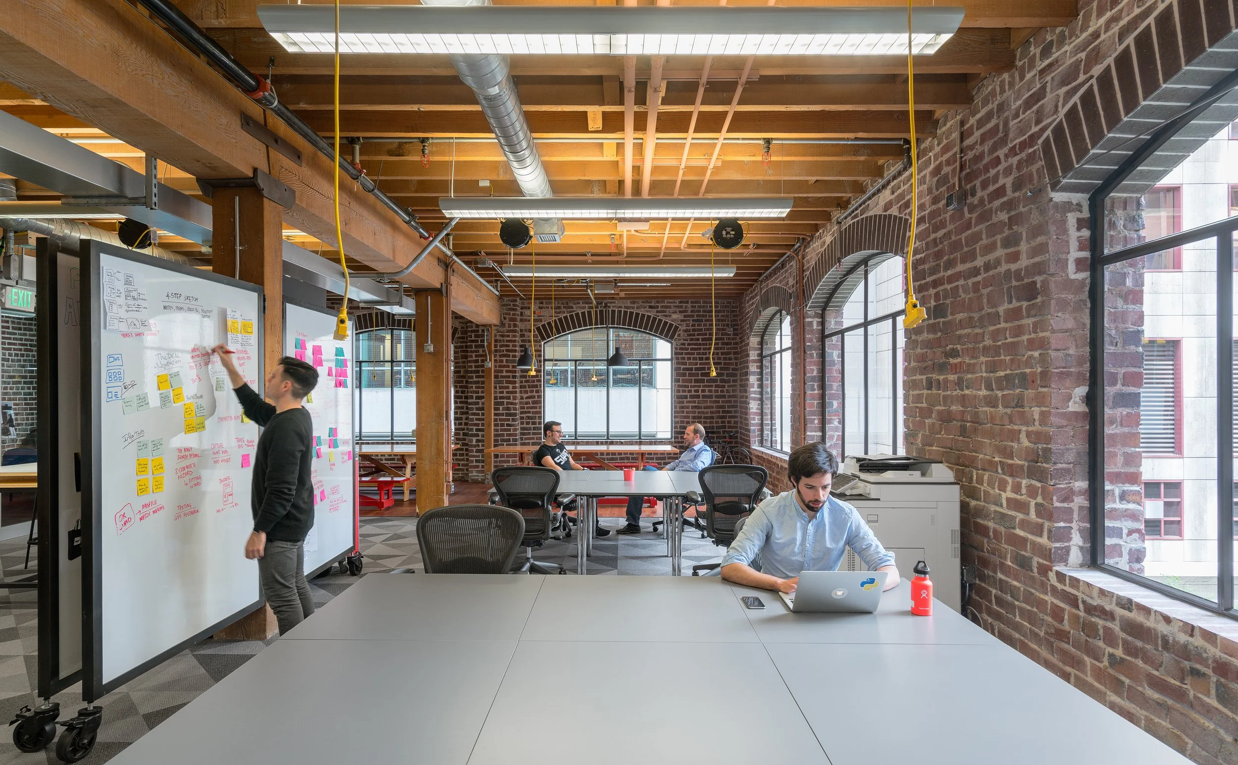 Office space with exposed brick walls, large windows, and wooden ceiling beams. Four people are working, one at a whiteboard with sticky notes and diagrams, two seated at a round table in the background, and one at a desk with a laptop and water bottle in the foreground.
