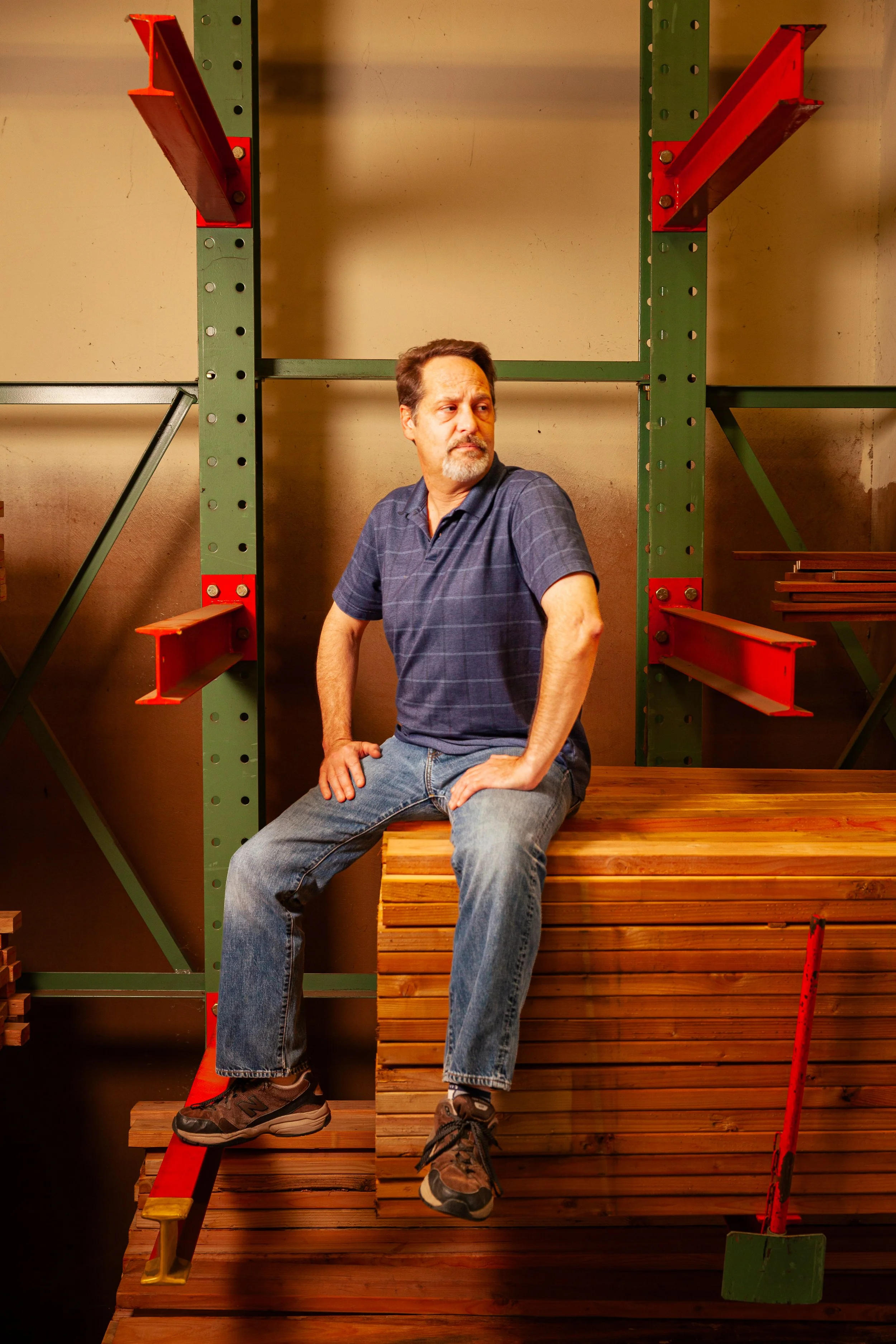 A middle-aged man with a beard, wearing a blue polo shirt and jeans, sitting on a wooden platform in a hardware store, surrounded by metal shelving and construction materials.