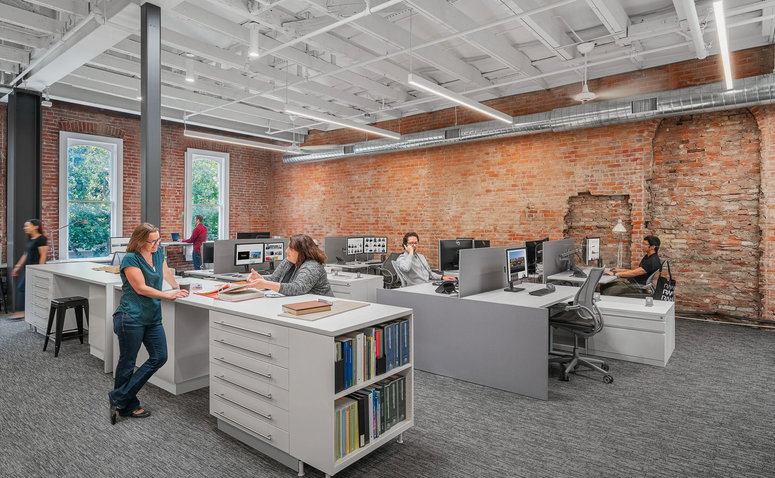 Open-plan office with exposed brick walls, large windows, modern desks with desktop computers, and several employees working at their stations. There's a woman with glasses and a blue polka-dot shirt talking to a colleague with brown hair. The office has a gray and white color scheme, a bookshelf, and visible ceiling ductwork.